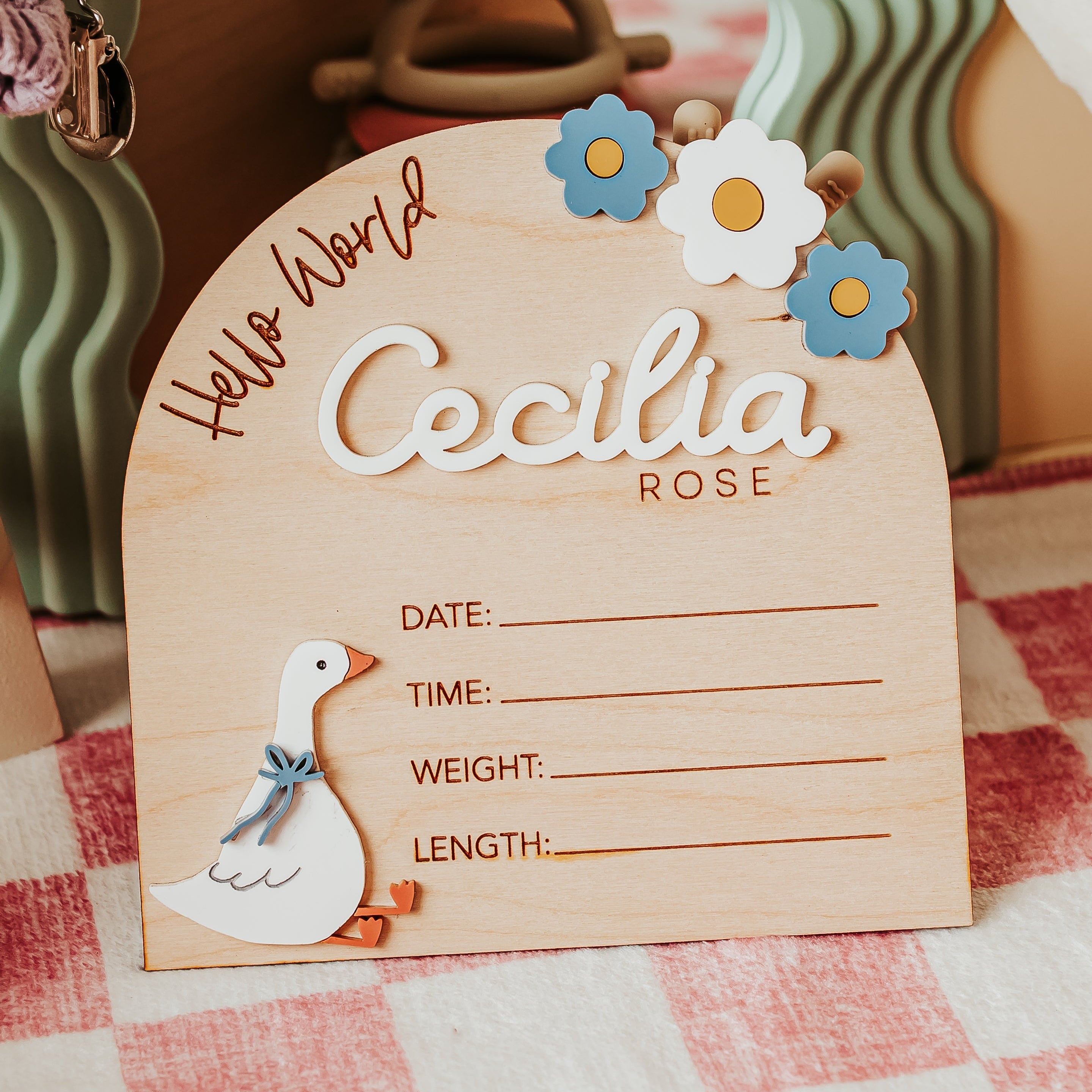 A personalized wooden baby birth stat sign with a silly goose with blue bow and blue and white flower design, placed on a pink and white checkered tablecloth. Sign reads 'Hello World', lists baby name Cecilia Rose and then lists date, time, weight, and length to write in baby's birth stats.
