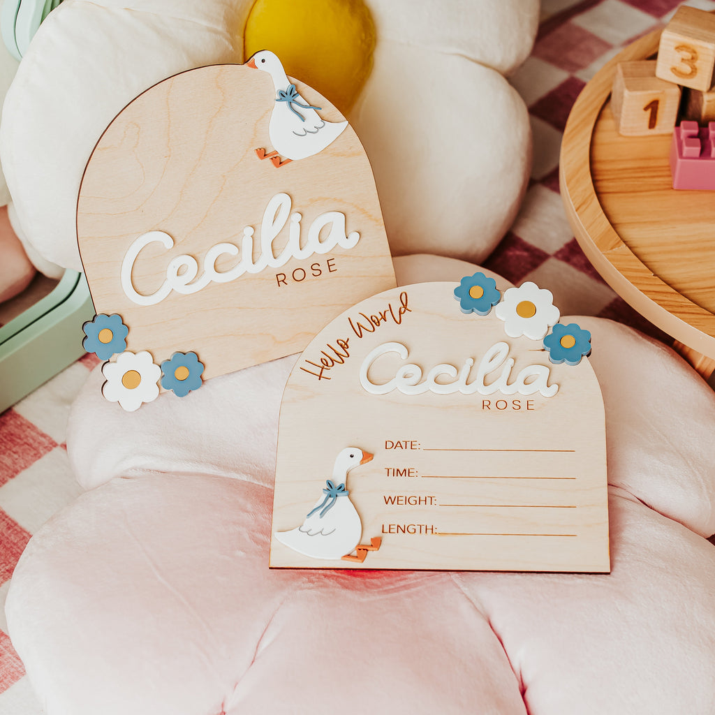 Two personalized wooden baby  name and baby birth stat signs with cute 3D acrylic goose and blue and white floral accents, displayed on a pink cushion over pink-and-white checkered blanket next to a wooden toy block tray.