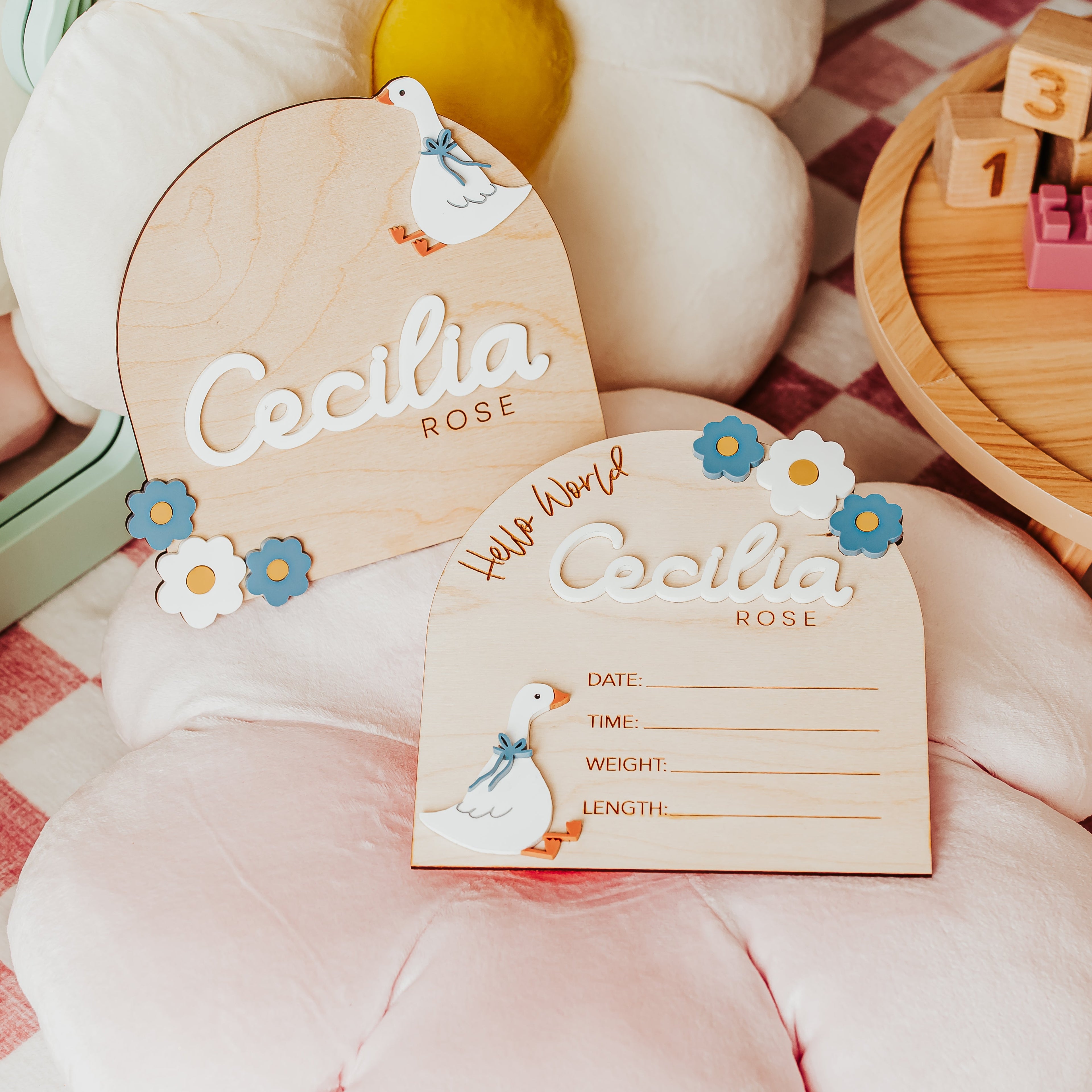 Two personalized wooden baby  name and baby birth stat signs with cute 3D acrylic goose and blue and white floral accents, displayed on a pink cushion over pink-and-white checkered blanket next to a wooden toy block tray.