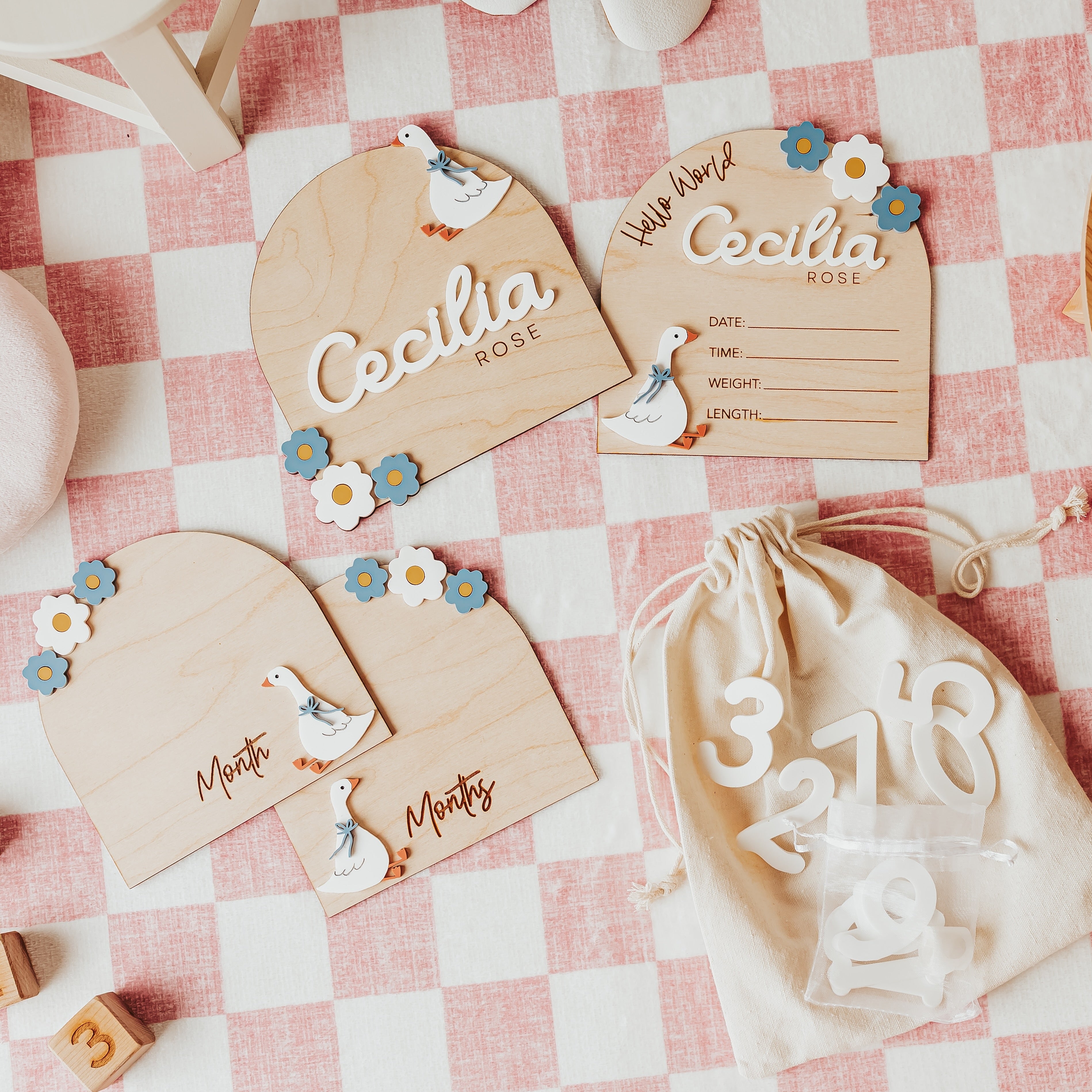 Personalized wooden baby baby announcement sets and matching drawstring bag laid out on a pink-and-white checkered blanket with baby toys around them. The cards are engraved with names, dates, and silly goose illustrations.