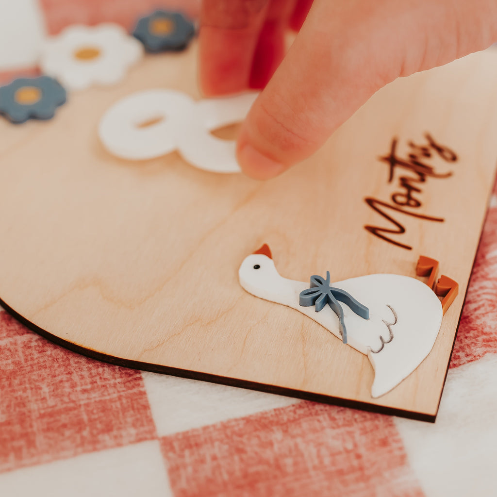 A close-up of a silly goose wooden monthly milestones or keepsake board with a hand placing a white die-cut duck sticker on it. The board has a natural light wood finish, an acrylic goose, white numbers, and small flower stickers, resting on a pink and white checkered fabric surface.