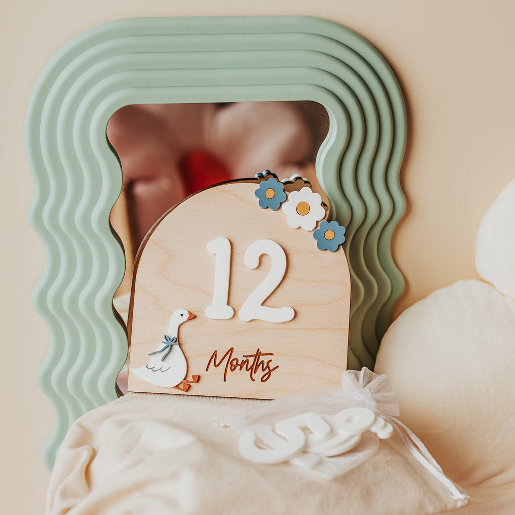 A wooden 12-month baby milestone plaque with white numbers and a swan illustration, styled on a cream fabric-covered chair in front of a wavy mint green mirror.