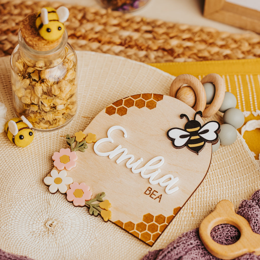 A styled flat-lay of baby teething toys and a wooden name plaque. The set includes a wooden honeycomb-patterned plaque engraved with 'Emilia BEA', a wooden bee teether, a wooden bead ring, a glass jar filled with dried oats topped with a bee figure, and a purple knit blanket.