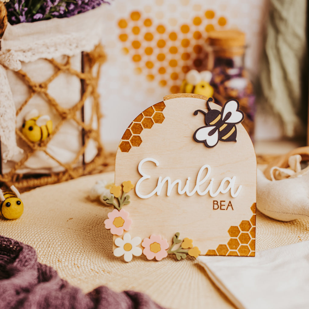 A personalized wooden plaque with an arched top, featuring the name 'Emilia' in white script and 'BEA' below it, decorated with honeycomb patterns, small flowers, and a wooden bee. The plaque is styled on a woven mat with a purple knit blanket, lavender, and bees in the background.