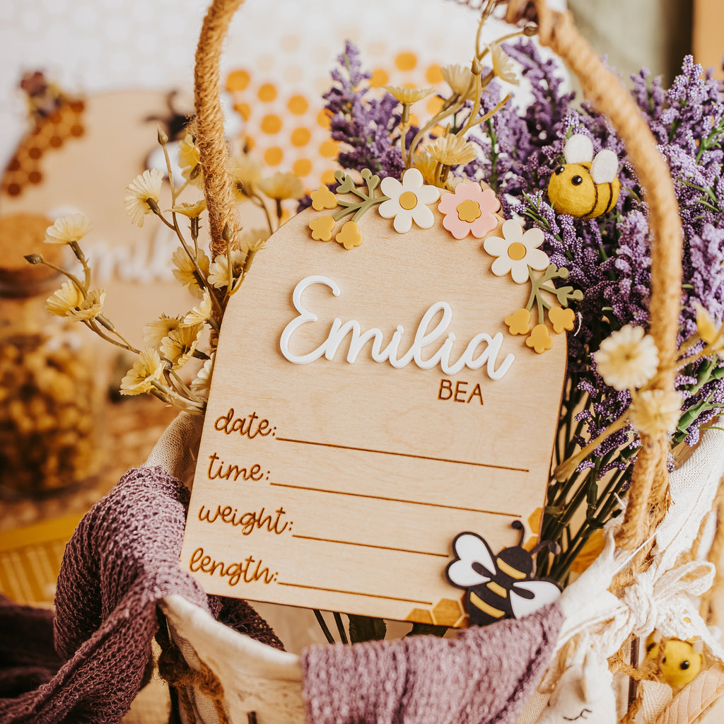 A rustic personalized baby birth stat with a wooden sign reading 'Emilia BEA' and fill-in lines, decorated with dried flowers, lavender, bumblebees, and a purple knit scarf.