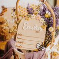 A rustic personalized baby birth stat with a wooden sign reading 'Emilia BEA' and fill-in lines, decorated with dried flowers, lavender, bumblebees, and a purple knit scarf.