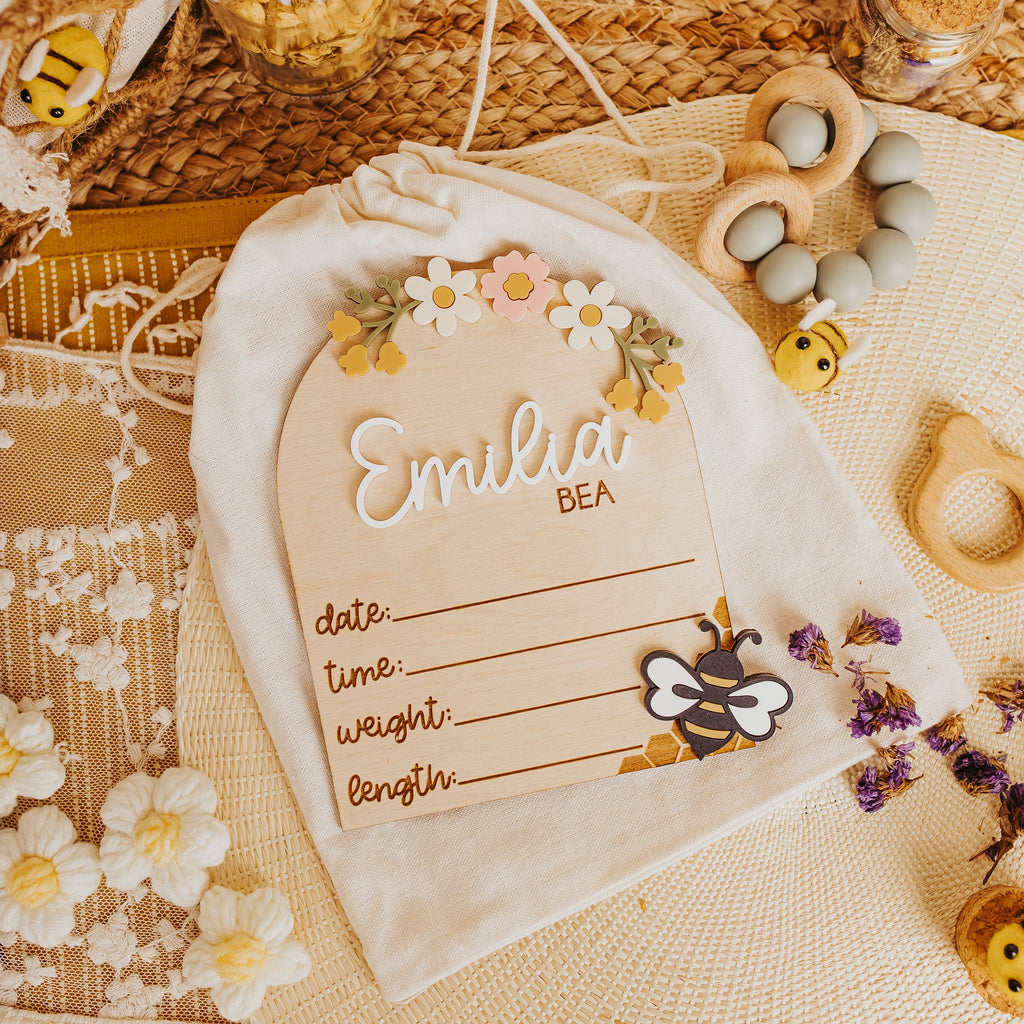 A cream-colored fabric baby keepsake bag with a drawstring closure. A baby birth stat lays on top  is personalized with the name 'Emilia BEA' decorated with a floral crown and a bee illustration.