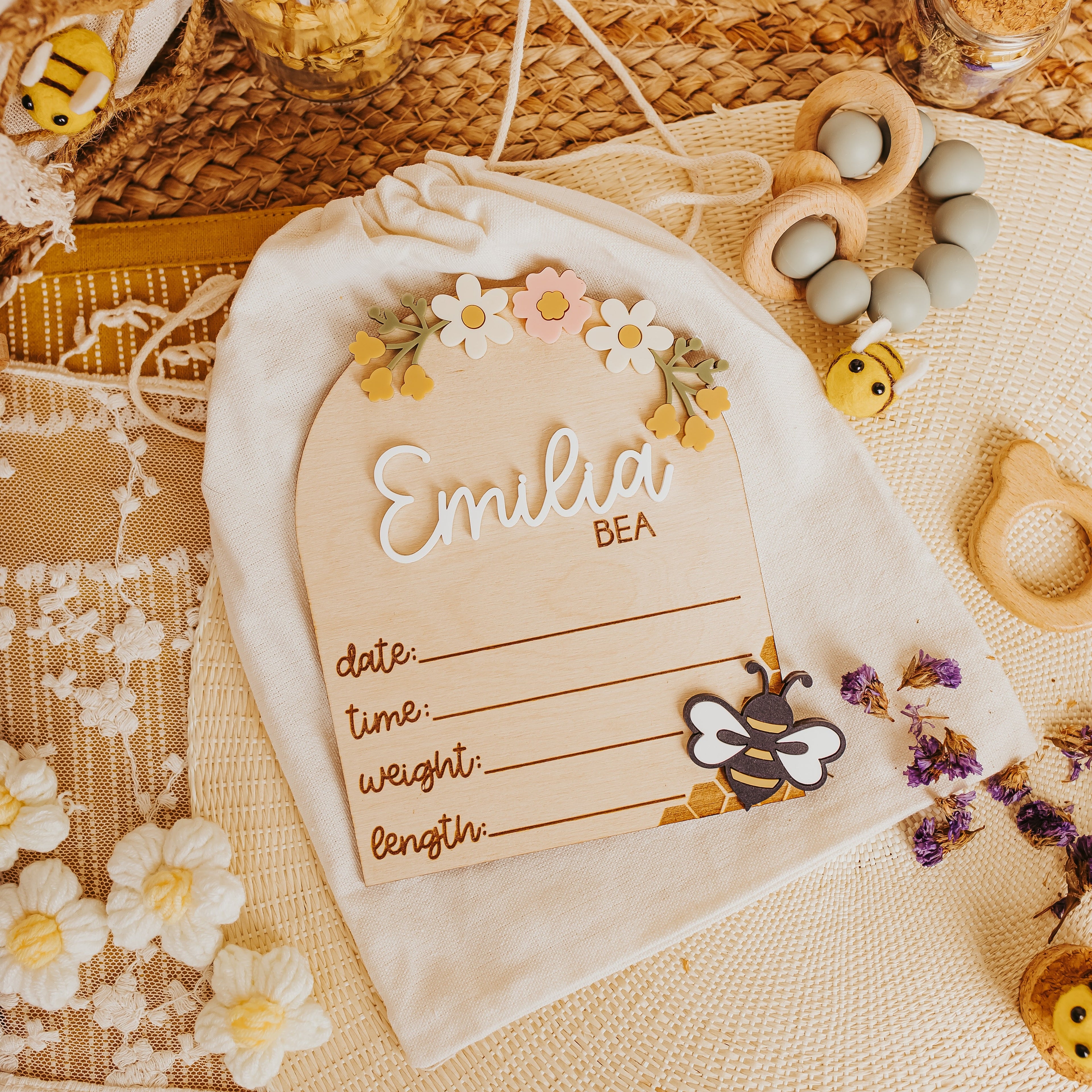 A cream-colored fabric baby keepsake bag with a drawstring closure. A baby birth stat lays on top  is personalized with the name 'Emilia BEA' decorated with a floral crown and a bee illustration.