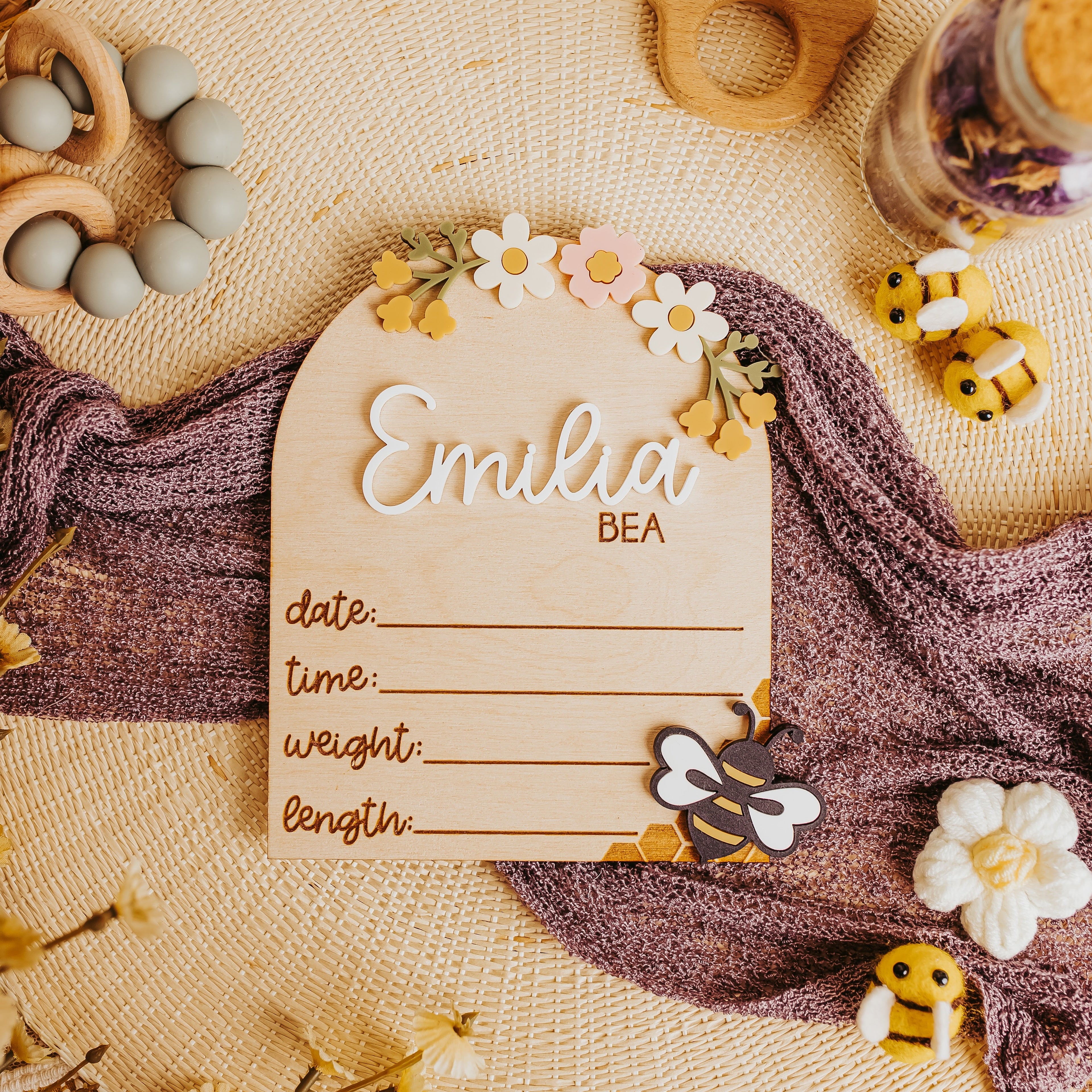 A personalized baby birth announcement card styled in a warm, bohemian flat-lay. The card is made of light natural wood with 'Emilia BEA' written in elegant cursive and block lettering, decorated with small white and yellow flowers.