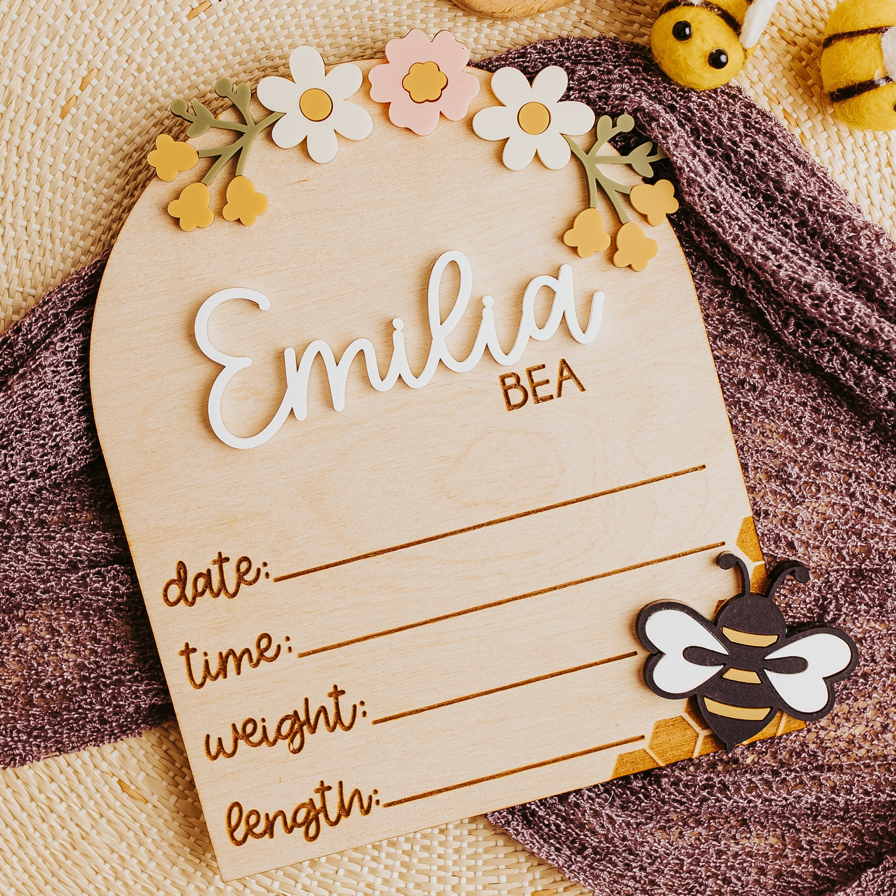 A personalized wooden baby birth stat sign with the name 'Emilia Bea' and fields for date, time, weight, and length, styled on a textured beige blanket with a purple knit scarf and bee-themed toys.
