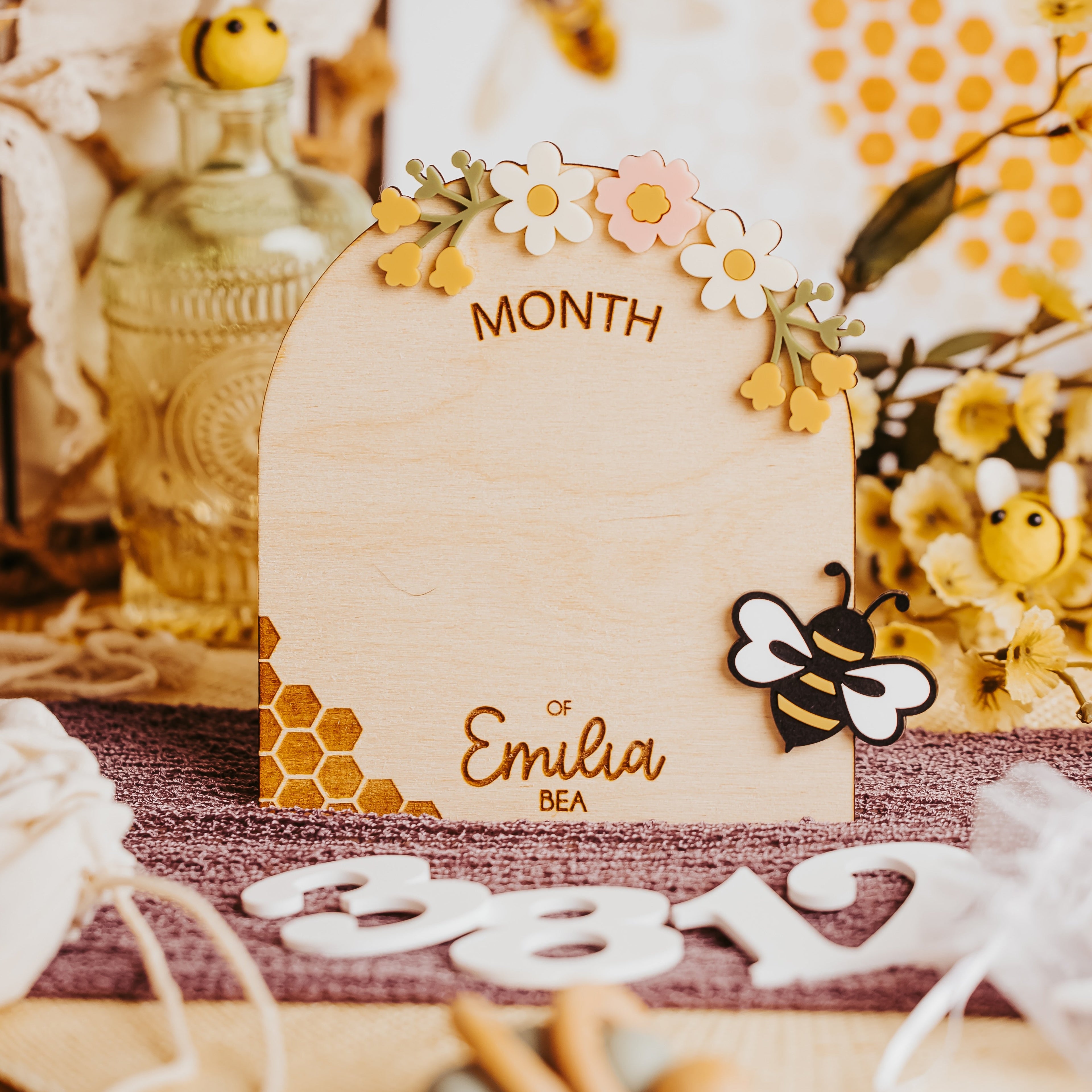 A wooden arch-shaped monthly milestone sign with floral and bee decorations, displayed on a rustic table with lavender, honeycomb props, and decorative numbers.