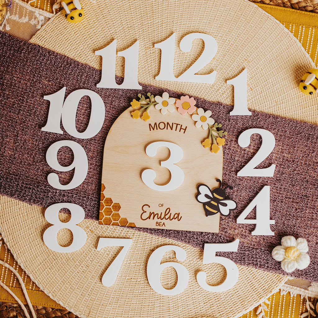 A round burlap with a central wooden month marker engraved with 'Month of Emilia' and engraved with a honey comb and acrylic flowers and a bee. The plaque is styled on a yellow woven mat with a brown ribbon.
