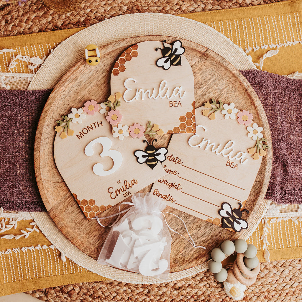 A styled flat-lay of a wooden baby milestone card, birth stat and name sign with a honeybee and floral designs, displayed on a woven mat with a purple ribbon and decorative accessories.