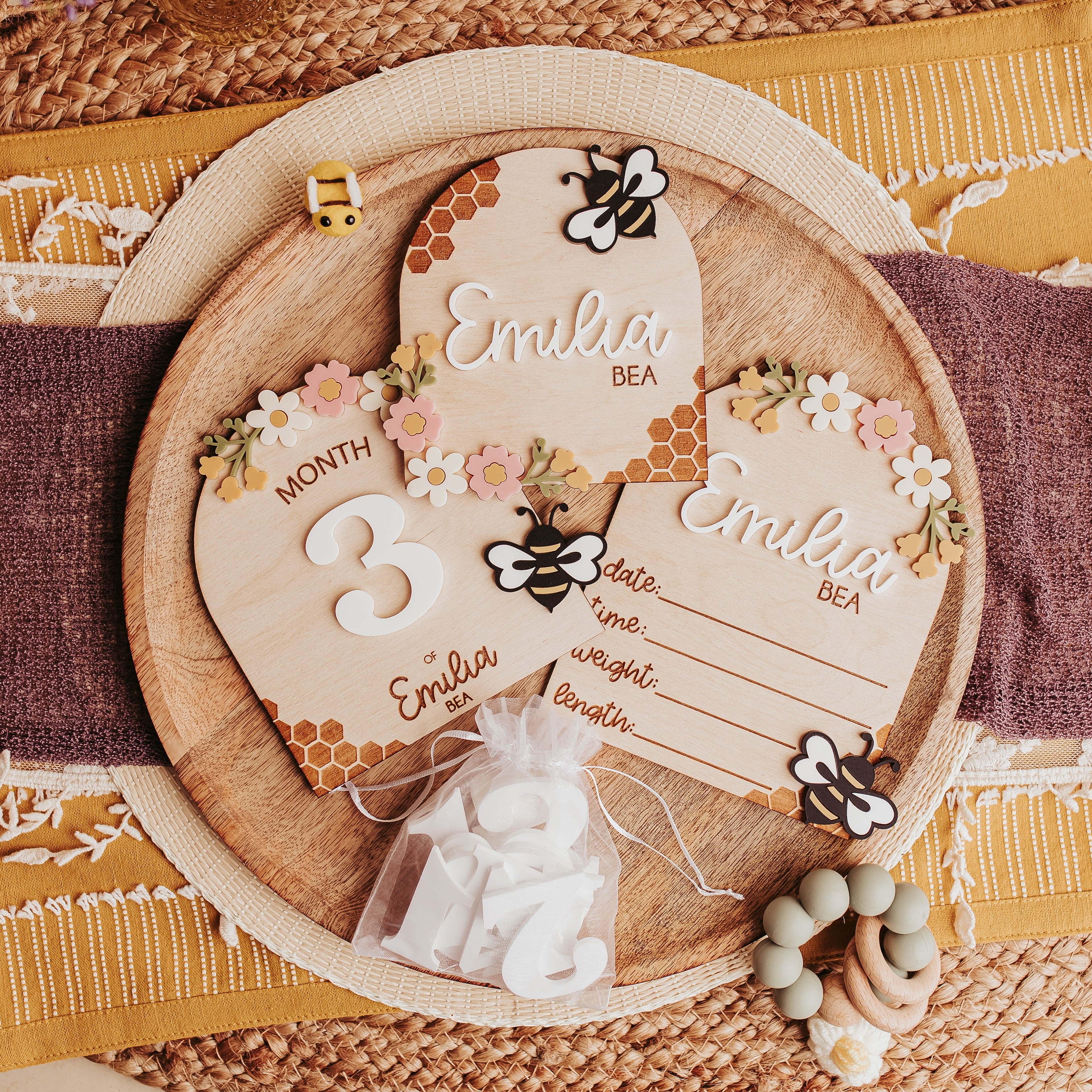 A styled flat-lay of a wooden baby milestone card, birth stat and name sign with a honeybee and floral designs, displayed on a woven mat with a purple ribbon and decorative accessories.