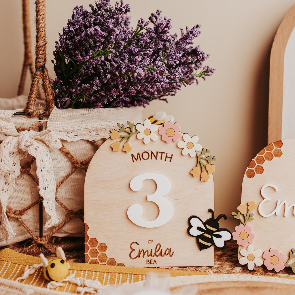A rustic wooden baby milestone card displaying the number 3 and the name 'Emilia', decorated with small flowers and a bee, styled with lavender in a basket on a beige background.