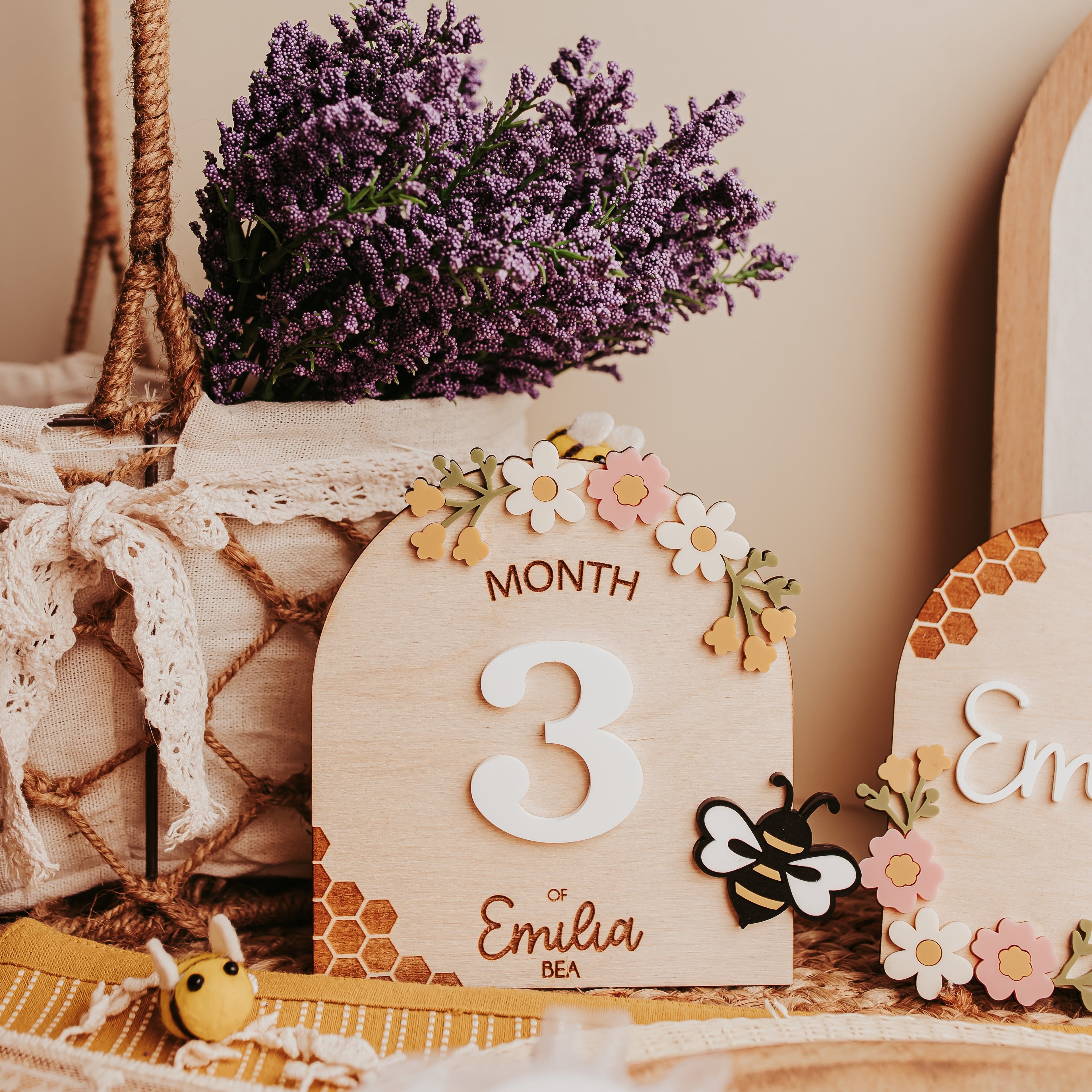 A rustic wooden baby milestone card displaying the number 3 and the name 'Emilia', decorated with small flowers and a bee, styled with lavender in a basket on a beige background.