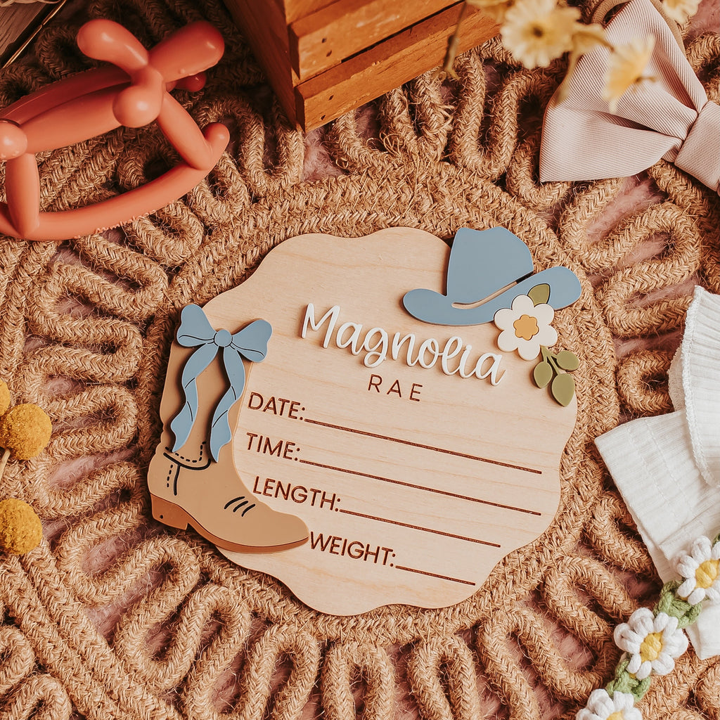 A wooden baby birth stat plaque reading 'Magnolia Rae' with lines for date, time, length, and weight, styled on a chunky woven rug with flowers, a wooden crate, and soft decorative props.