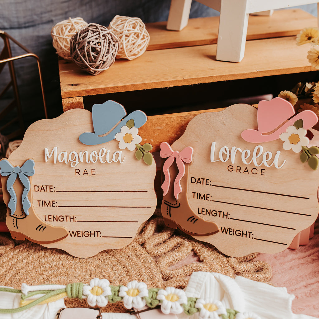Two wooden baby birth signs with cowboy hat and flower decorations, styled on a rustic wooden surface with woven textiles and floral decor.