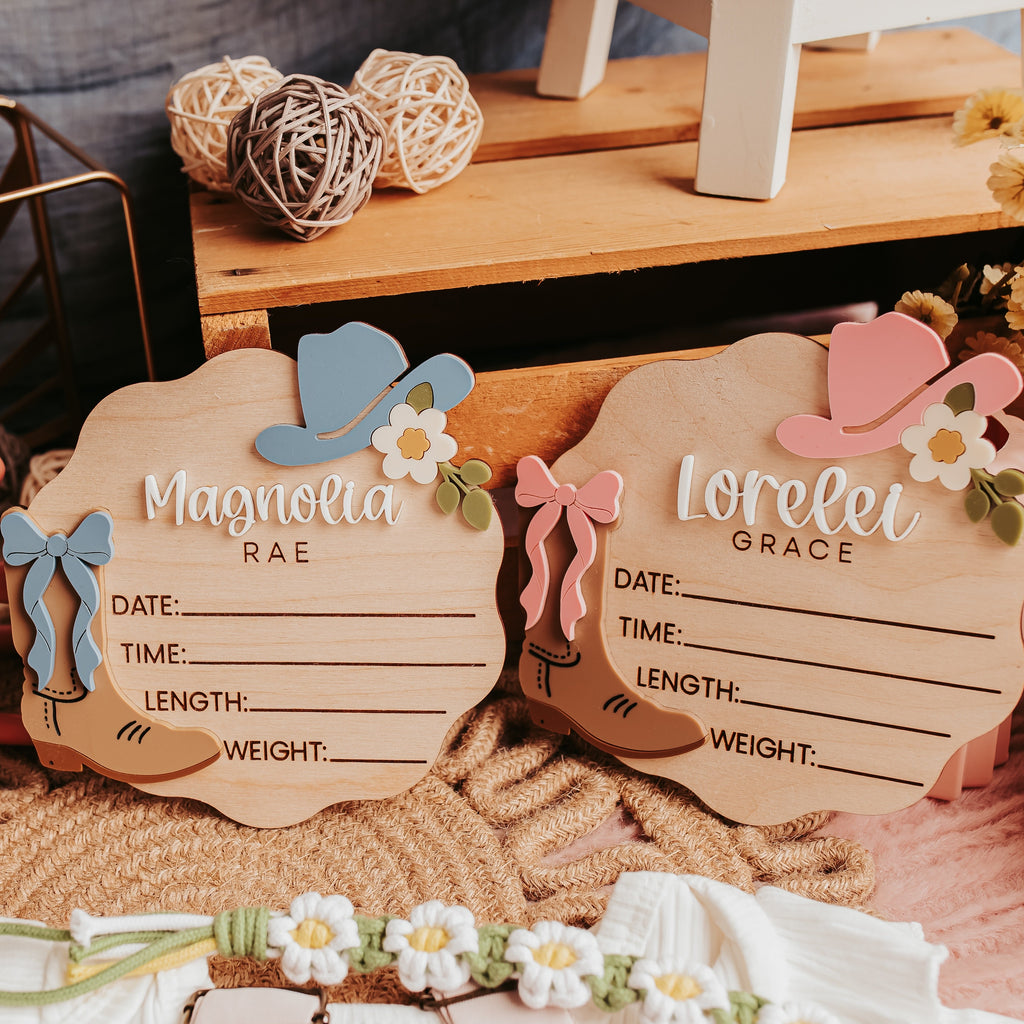Two wooden baby name signs with cowboy hat and flower decorations, styled on a rustic wooden surface with woven textiles and floral decor.