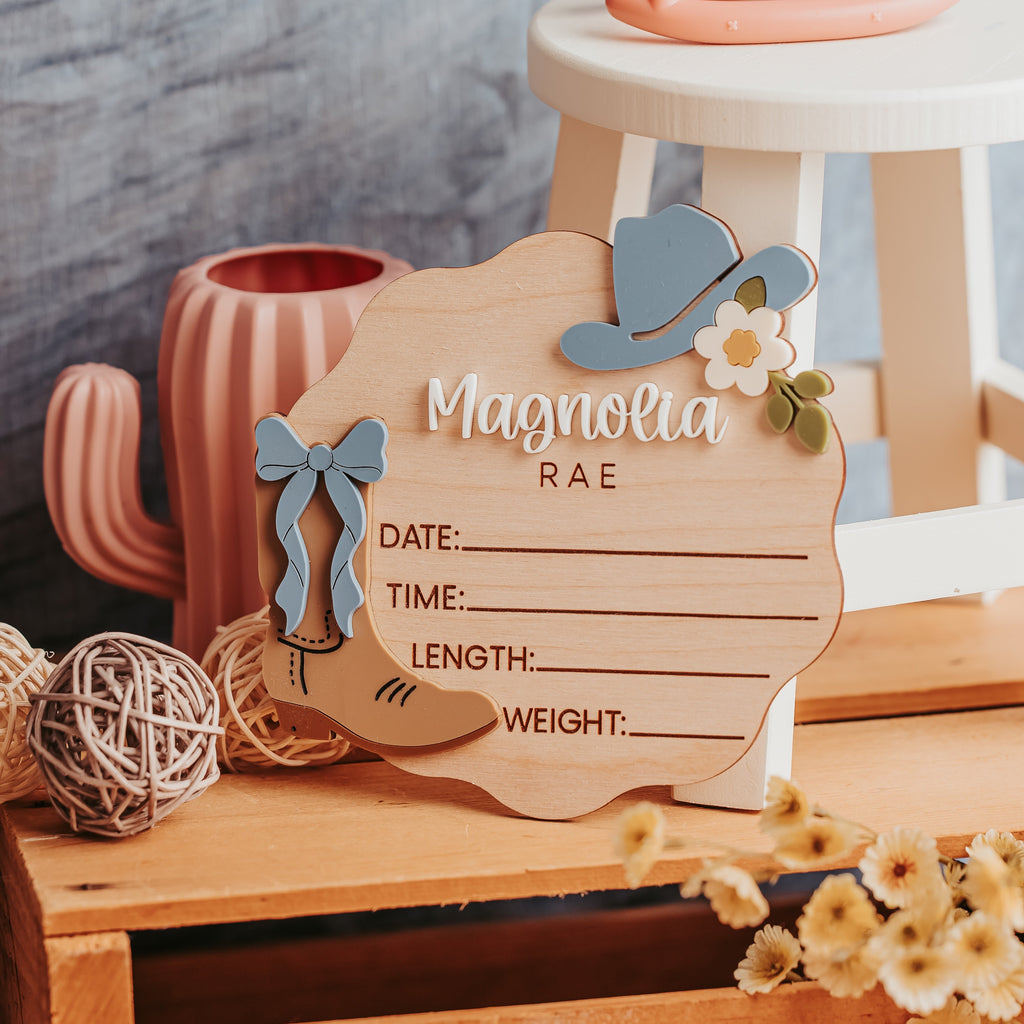 A wooden baby birth stat with a western theme, featuring a cowboy hat, bow, and floral accents, styled on a rustic wooden crate with decorative props.