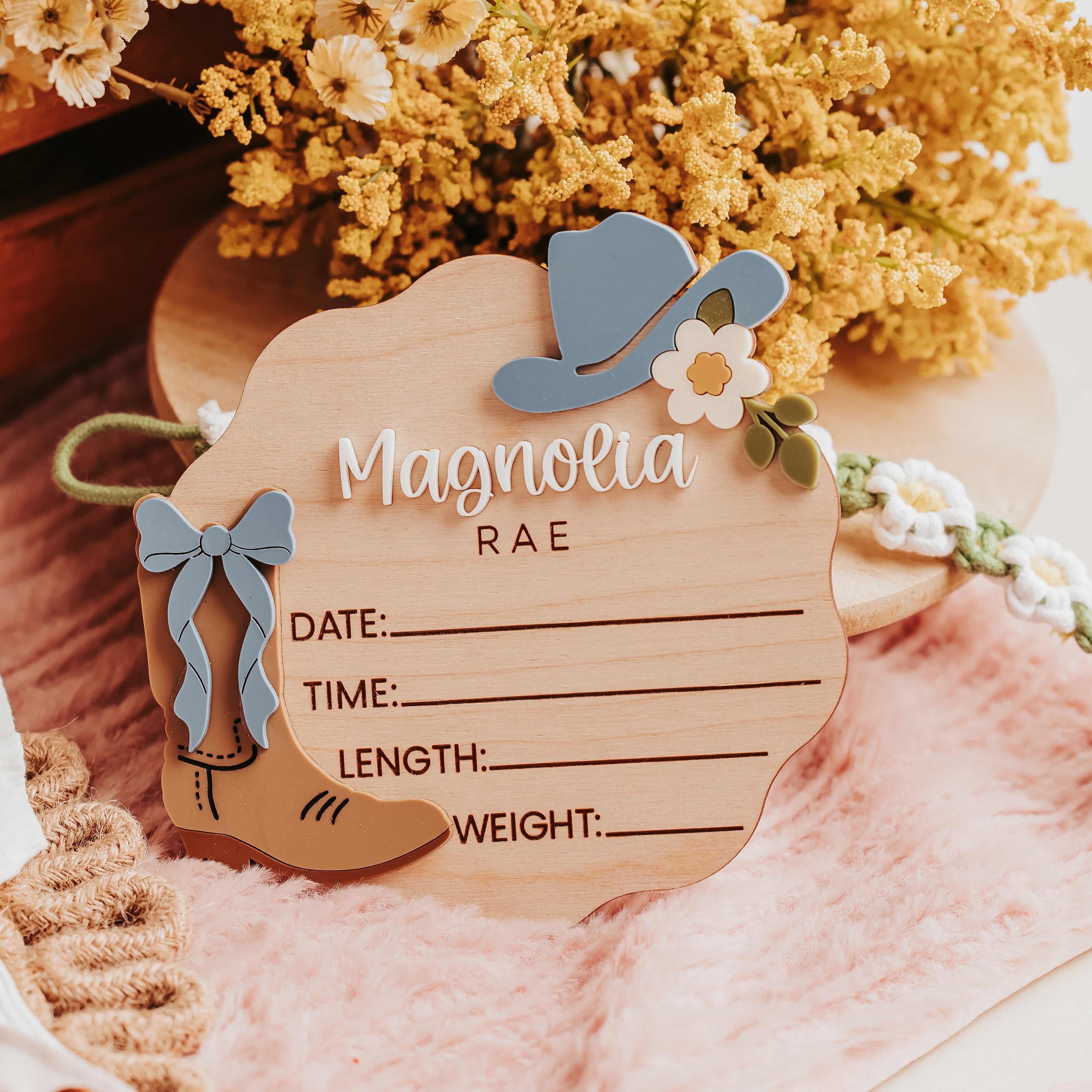 A wooden baby birth stat sign with a western cowgirl theme, featuring a blue hat, bow, flower, and boot cutout. The name 'Magnolia' and 'Rae' are printed on the wooden board, which is styled with dried yellow flowers, a pink blanket, and a white knit blanket.