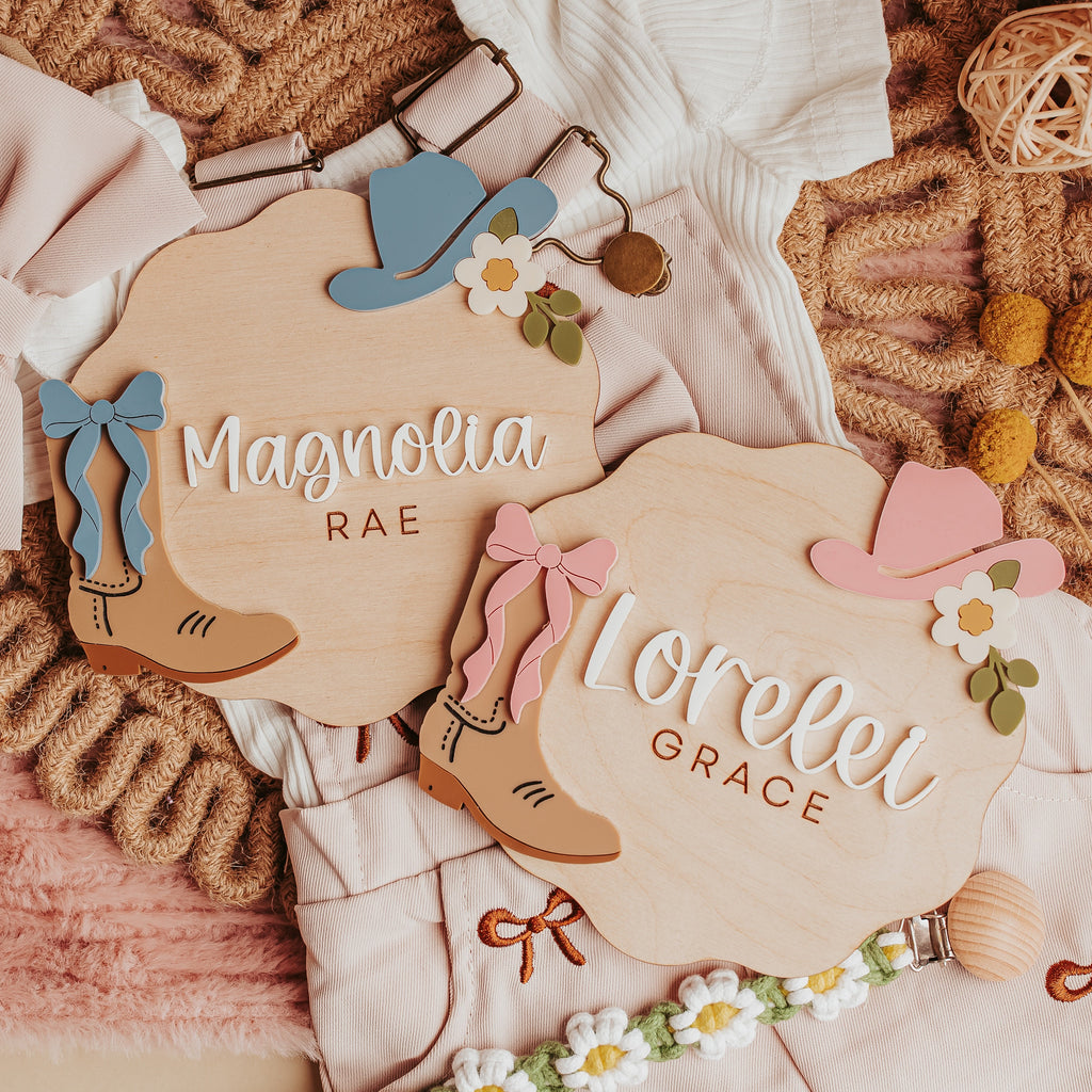 Two personalized wooden baby name signs with cowboy boot and hat illustrations, styled on a pink textured blanket with baby clothes, a bow, yarn balls, and daisy garland.