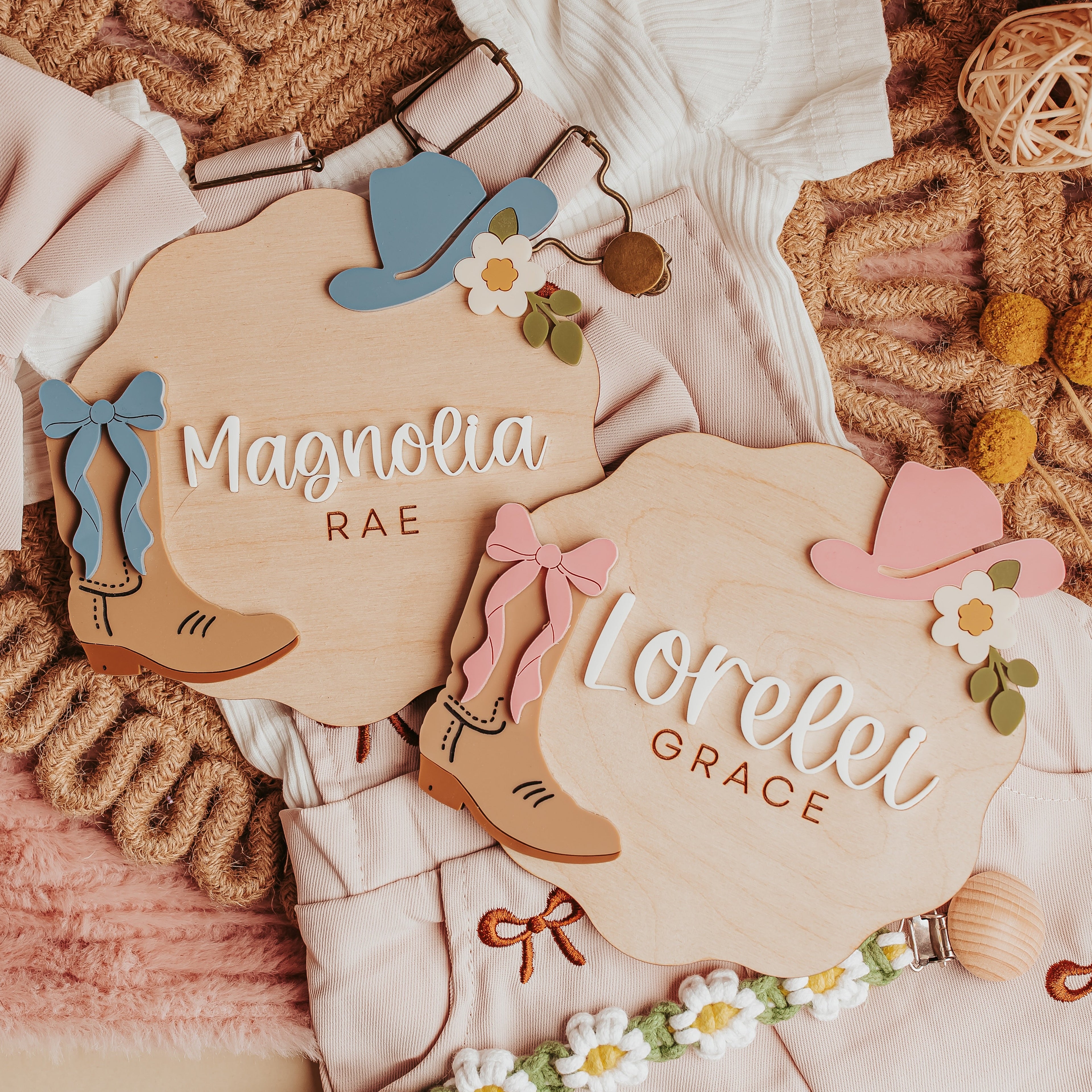 Two personalized wooden baby name signs with cowboy boot and hat illustrations, styled on a pink textured blanket with baby clothes, a bow, yarn balls, and daisy garland.