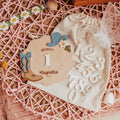 A styled flat-lay of a wooden first birthday milestone card reading 'Month 1 of Magnolia Rae' with a cowboy hat and boots design, placed on a cream fabric drawstring bag with white number cutouts, on a pink woven mat with daisy garland and dried flowers.