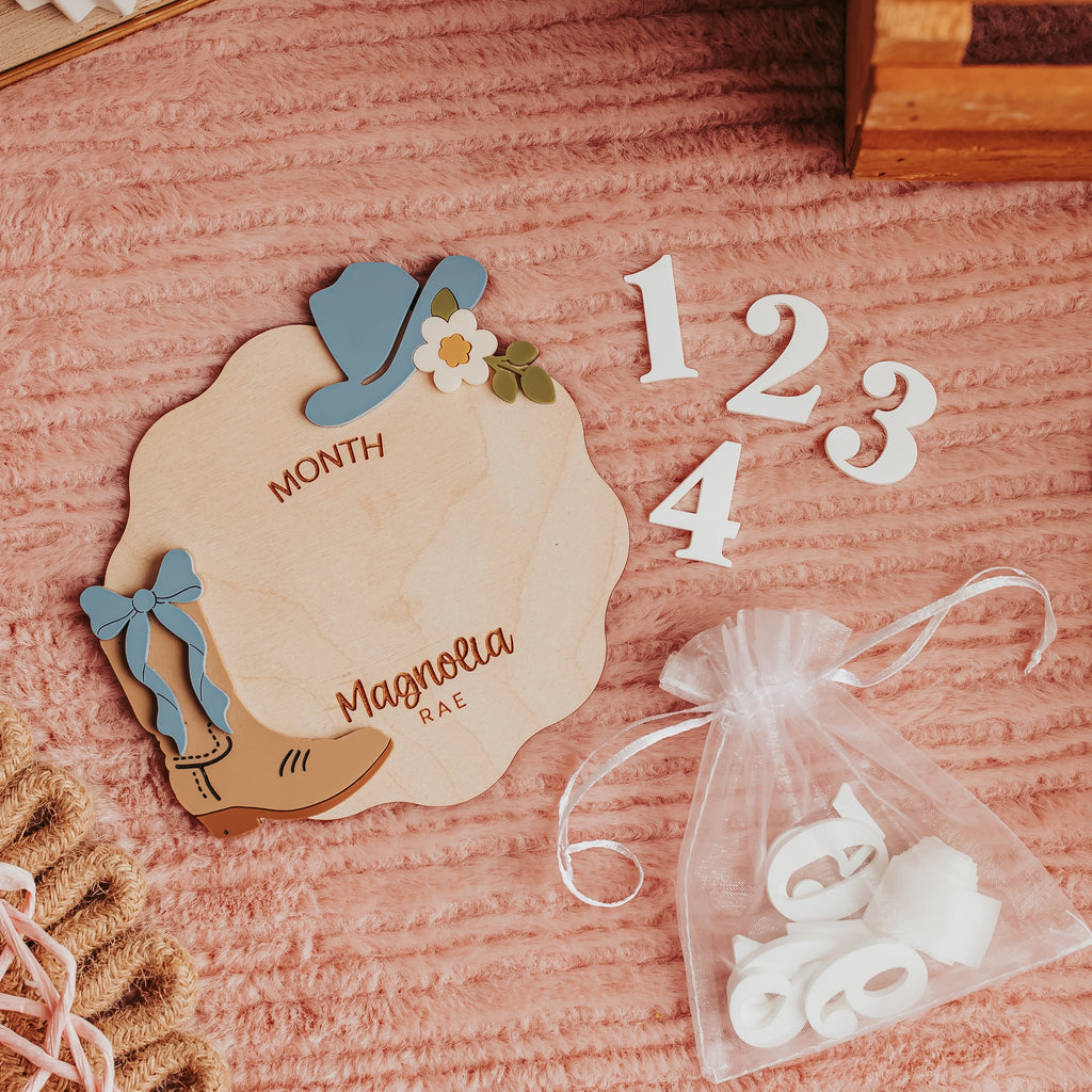 A styled flat-lay of a wooden milestone month and name card with cowgirl-themed decorations, white numbers 1, 2, 3, and 4, and a small clear bag of white letter cutouts on a pink textured fabric background.