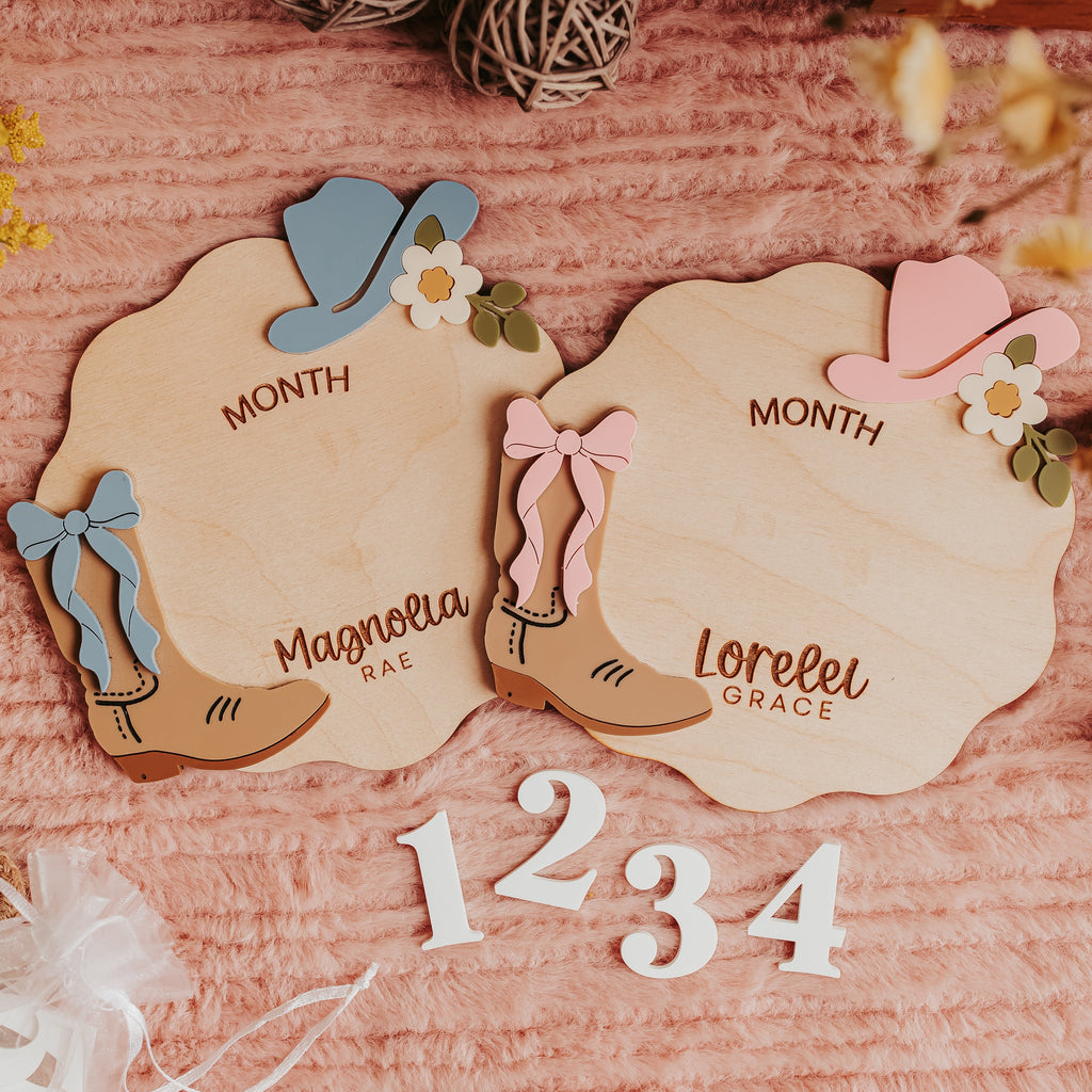 Two scalloped wooden baby monthly milestone plaques with cowboy boot illustrations, displayed on a pink textured fabric background with dried flowers and white numbers 1, 2, 3, 4.