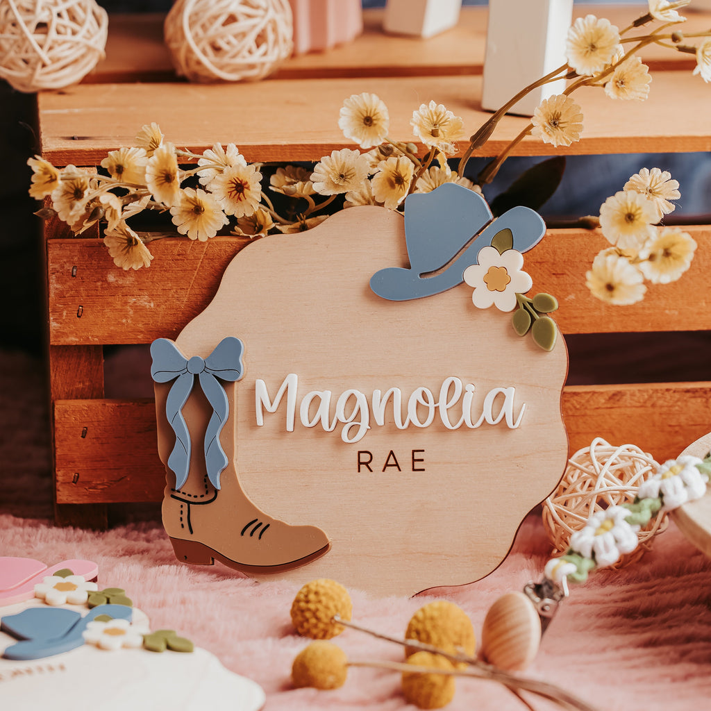 A rustic wooden personalized baby name sign reading 'Magnolia RAE' styled with a blue cowboy hat, blue bow, tan cowboy boot, and small white flowers on a pink fabric surface.