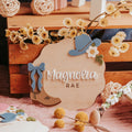 A rustic wooden personalized baby name sign reading 'Magnolia RAE' styled with a blue cowboy hat, blue bow, tan cowboy boot, and small white flowers on a pink fabric surface.