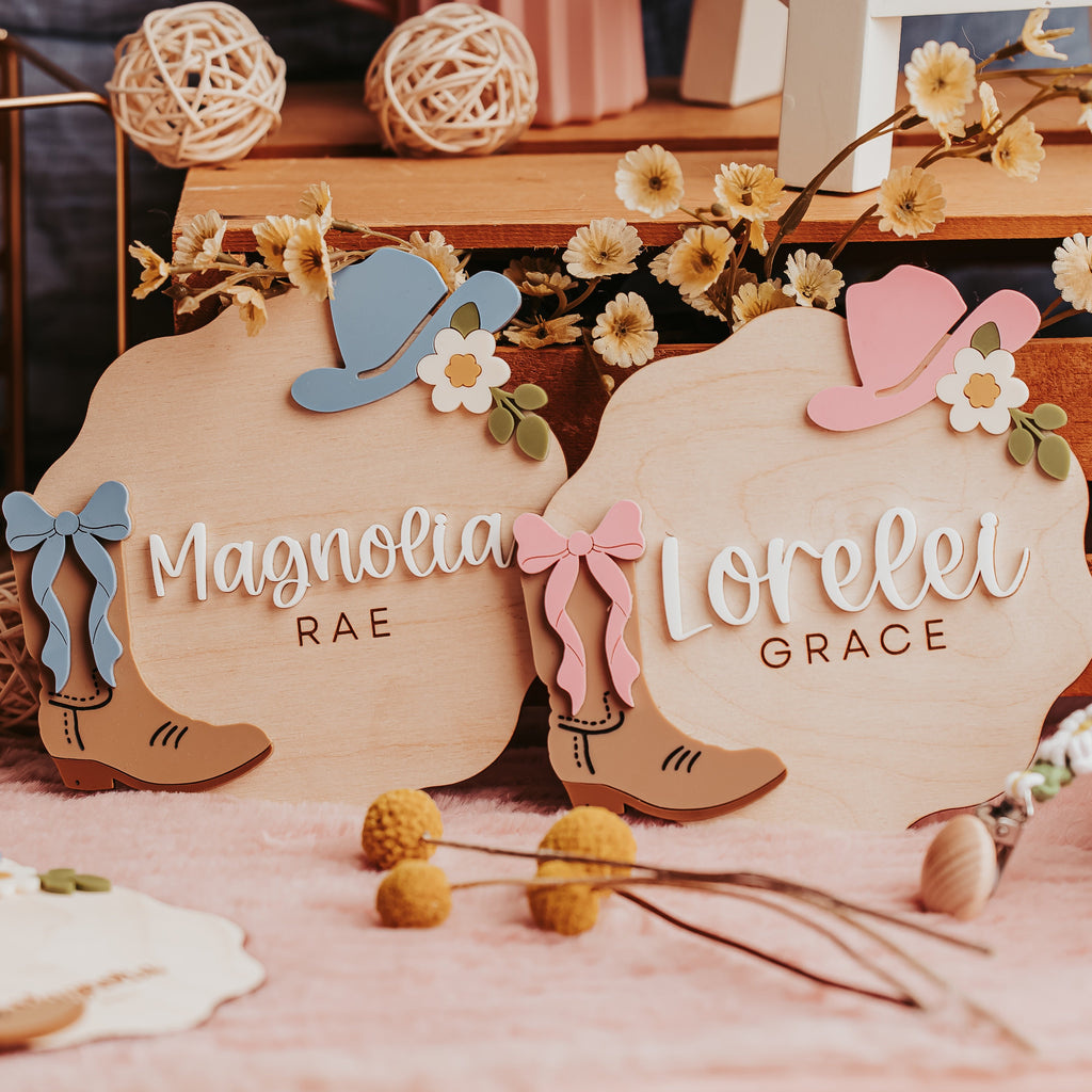 Two personalized wooden baby name signs with cowgirl boot and hat designs, styled on a pink fabric surface with dried flowers and rustic decor in the background.