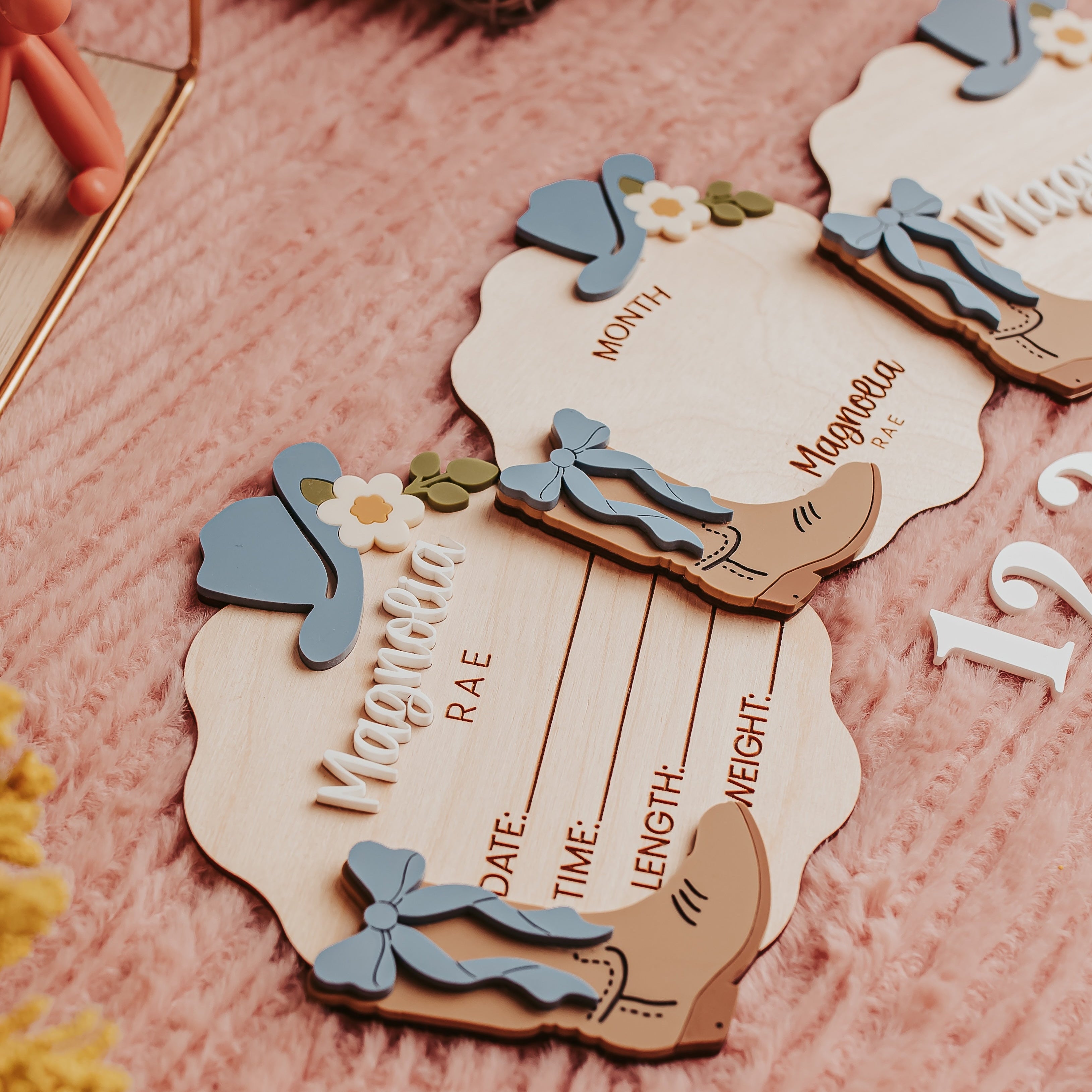 A close-up of decorative wooden baby announcement signs cards for a baby, styled on a pink textured blanket with yarn balls and dried flowers.