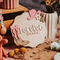 A personalized wooden baby name sign reading 'Lorelei Grace' styled in a cozy boho nursery setting with pink bows, a cactus-shaped vase, and warm rustic props.