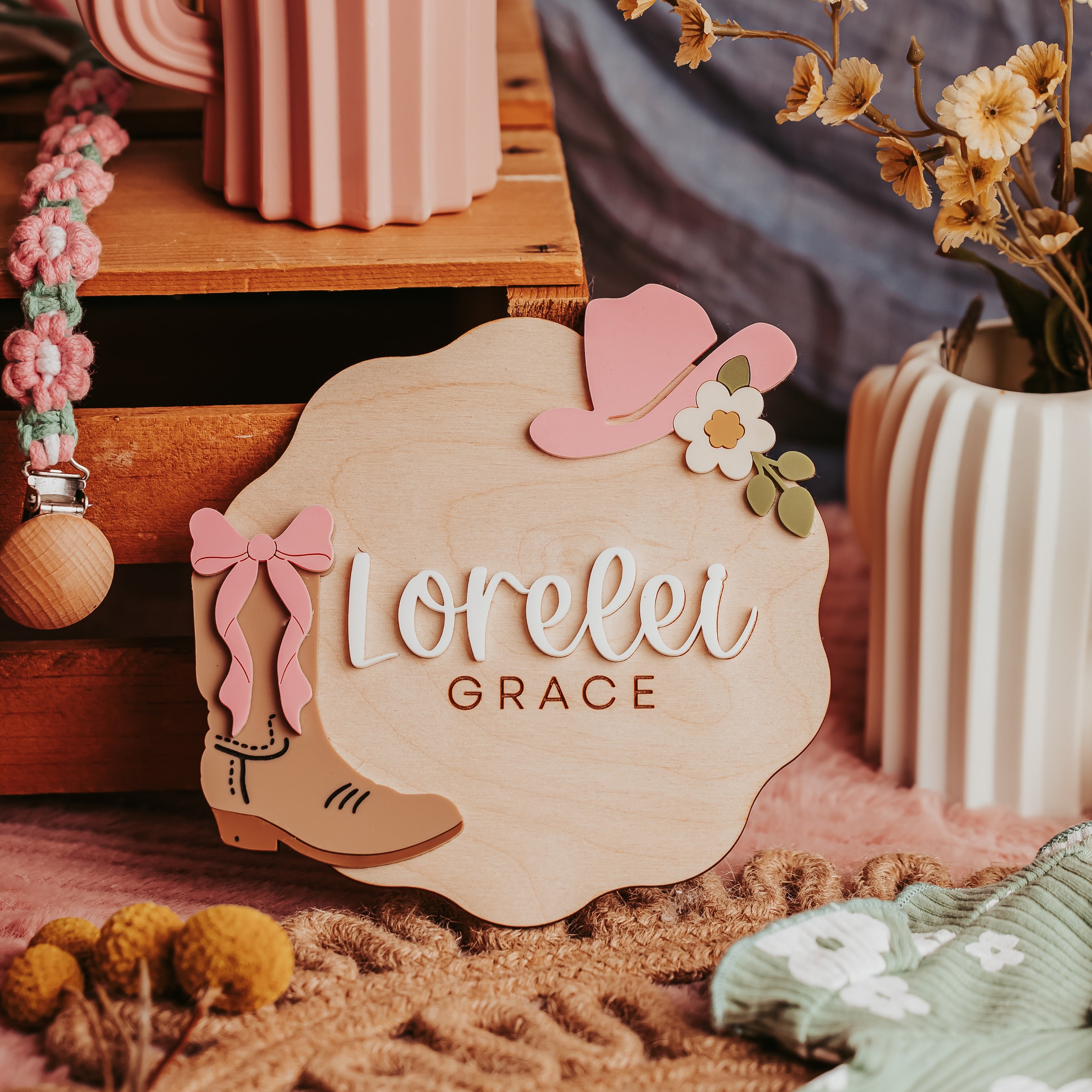 A personalized wooden baby name announcement sign with the name 'Lorelei Grace' in white script, decorated with a pink cowboy hat, pink bow, brown boot, and a small white flower. The sign is styled in a rustic boho setting with a pink cactus-shaped vase, dried flowers, and a brown knit blanket.