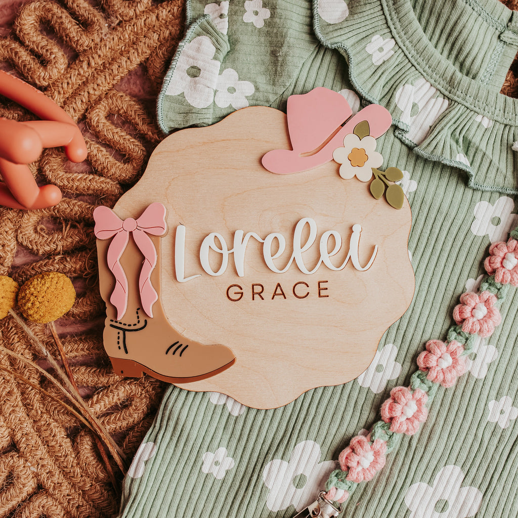A styled flat-lay of a baby girl's ribbed sage green romper with white floral prints, featuring scalloped wooden baby name announcement sign featuring a pink cowboy hat and engraved with 'Lorelei GRACE' and decorated with a pink cowboy boot and bow.