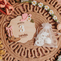 A styled flat-lay of a wooden baby milestone plaque engraved with 'MONTH 3 Lorelei GRACE', surrounded by baby accessories including a small clear bag of white footprints, a pink bow, and a beaded necklace on a woven placemat.