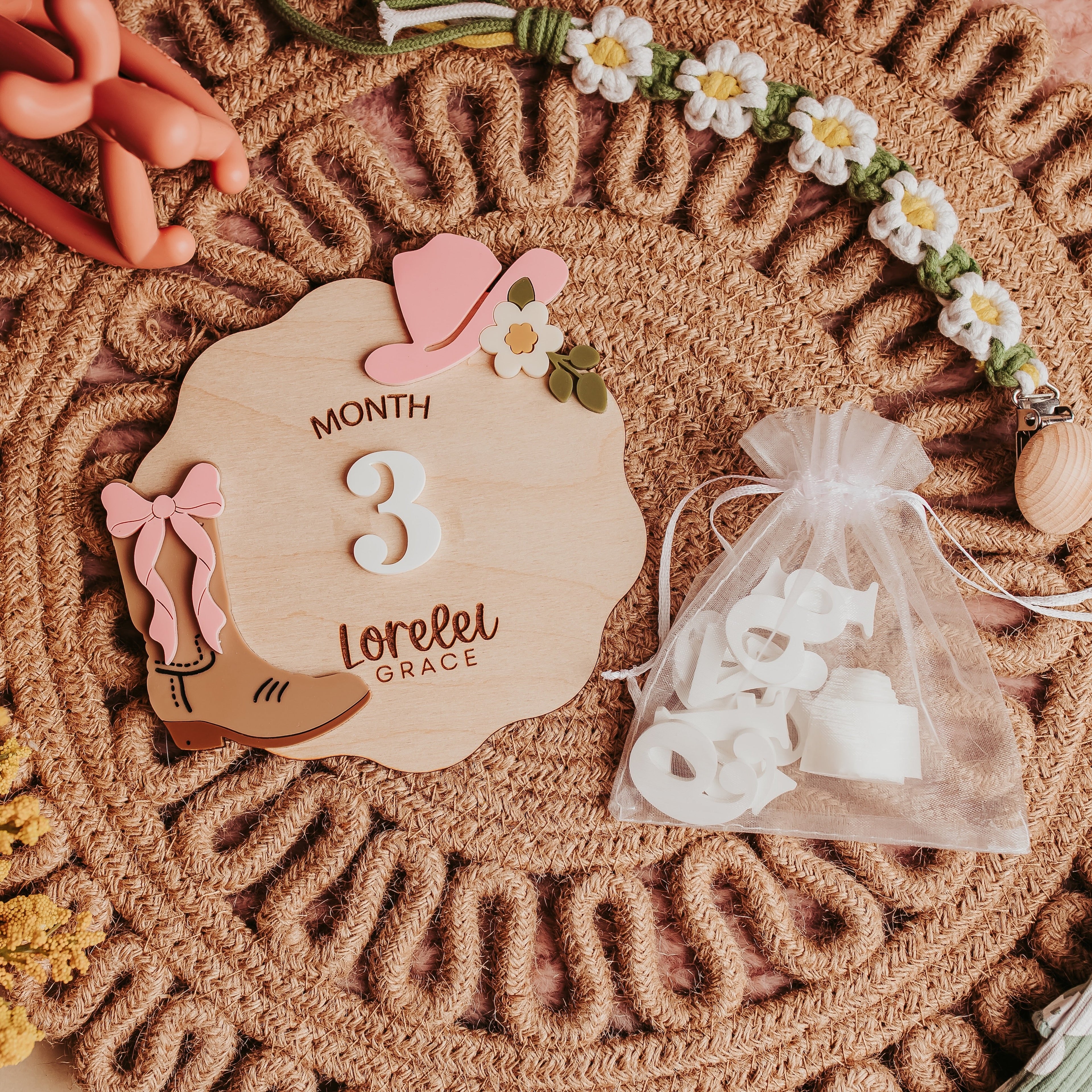 A styled flat-lay of a wooden baby milestone plaque engraved with 'MONTH 3 Lorelei GRACE', surrounded by baby accessories including a small clear bag of white footprints, a pink bow, and a beaded necklace on a woven placemat.