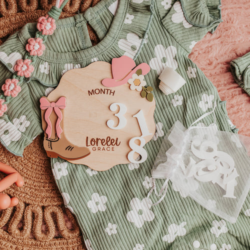 A styled flat-lay of a sage green ribbed baby romper with white floral prints, accompanied by a matching headband, a wooden milestone sign, and baby accessories on a pink textured blanket.