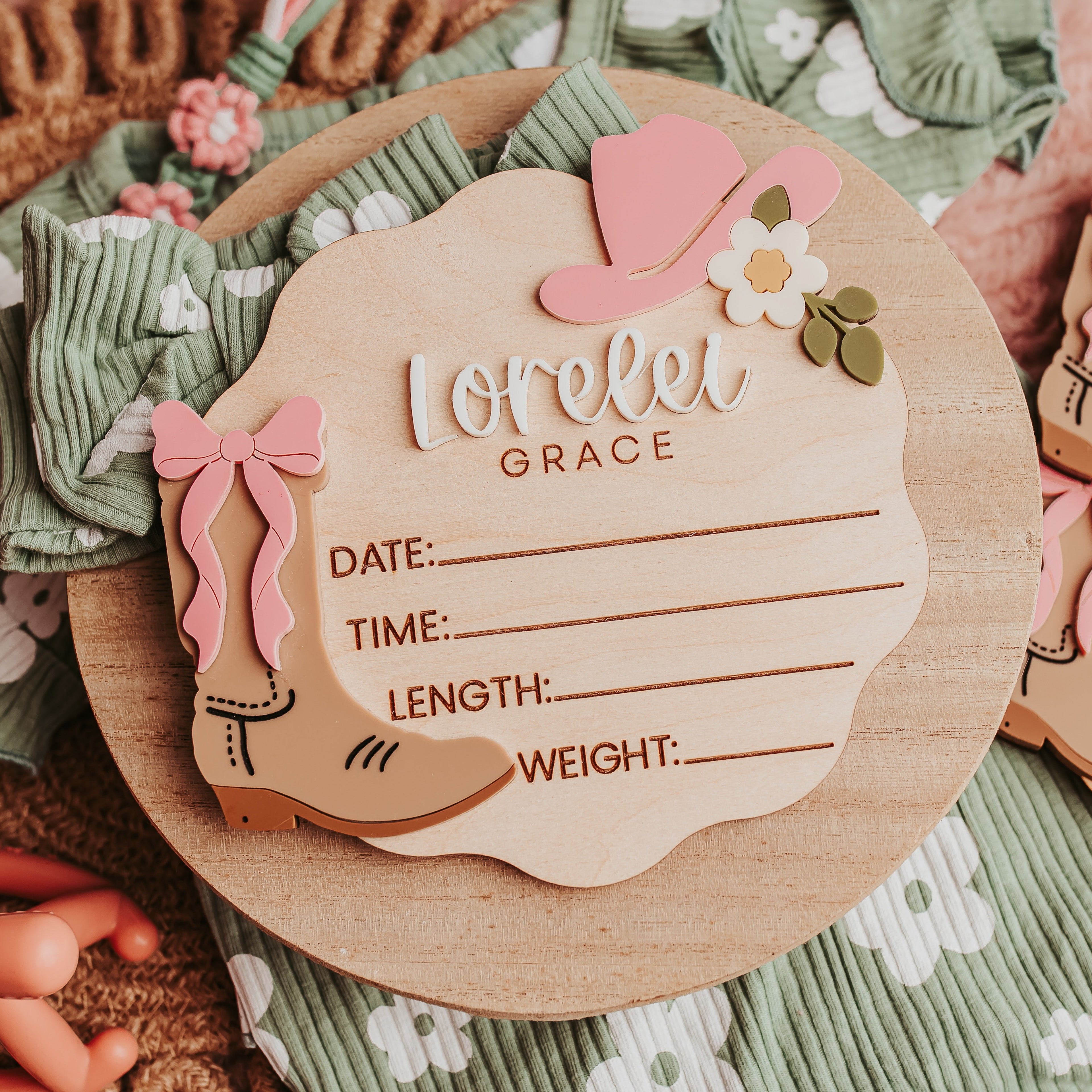 A wooden baby birth stat with a western theme, featuring a wooden boot, pink hat, bow, and fill-in lines for date, time, length, weight, and name. Styled on a green patterned fabric with other matching cards and props. Great for fresh 48 photoshoots.