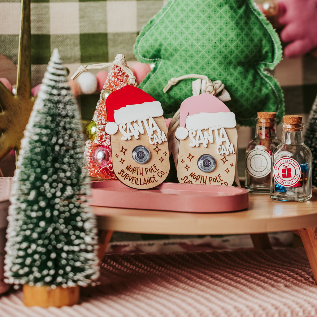 A styled holiday tabletop scene with two small faux Santa Cam ornaments s in the center, a green fabric Christmas tree behind them, a small frosted tree on the left, and decorative bottles on the right. 