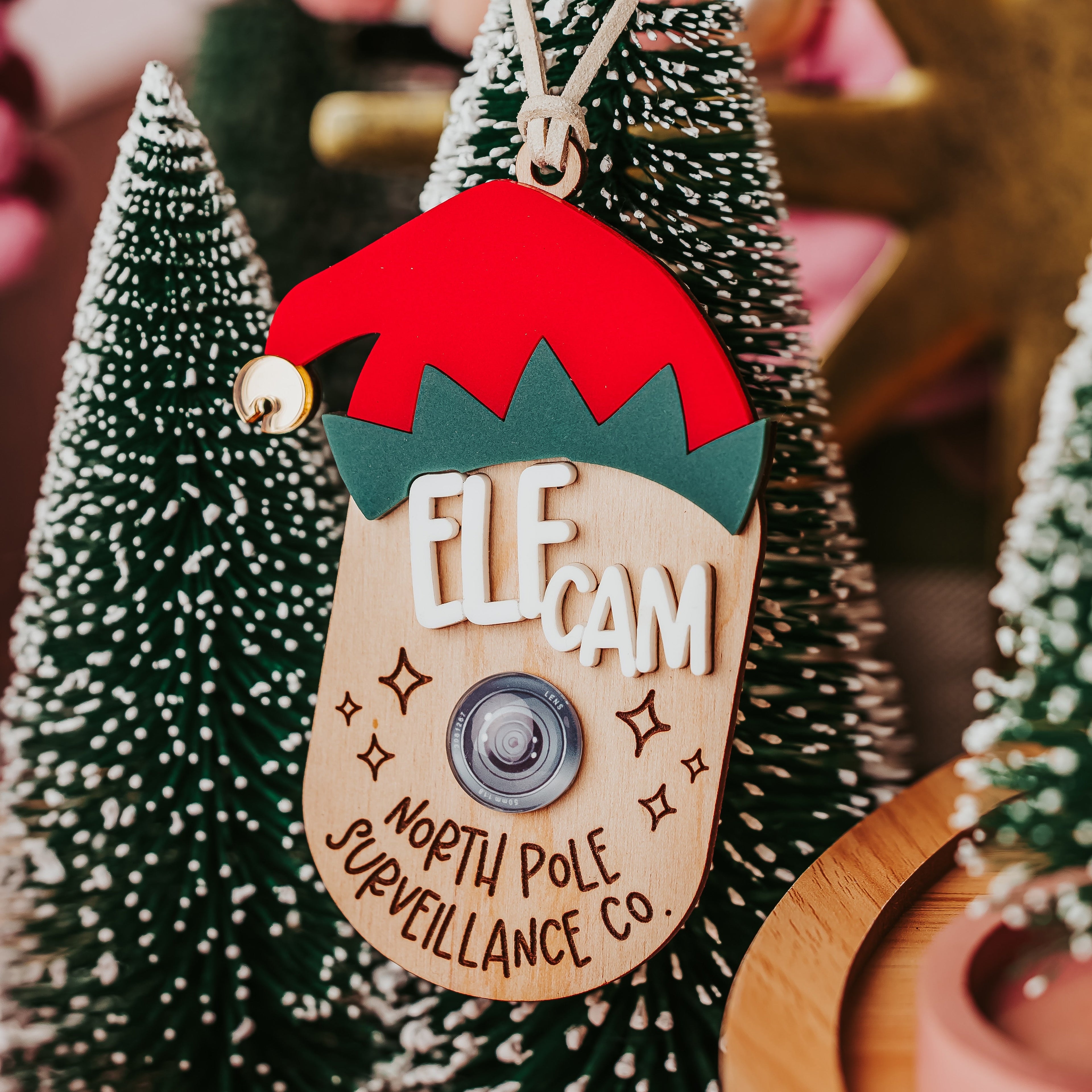 A Christmas wooden faux elf cam ornament shaped like an elf head wearing a red Santa hat and green collar, with a faux  camera button at the center. The ornament reads 'ELF CAM' and 'NORTH POLE SURVEILLANCE CO.'
