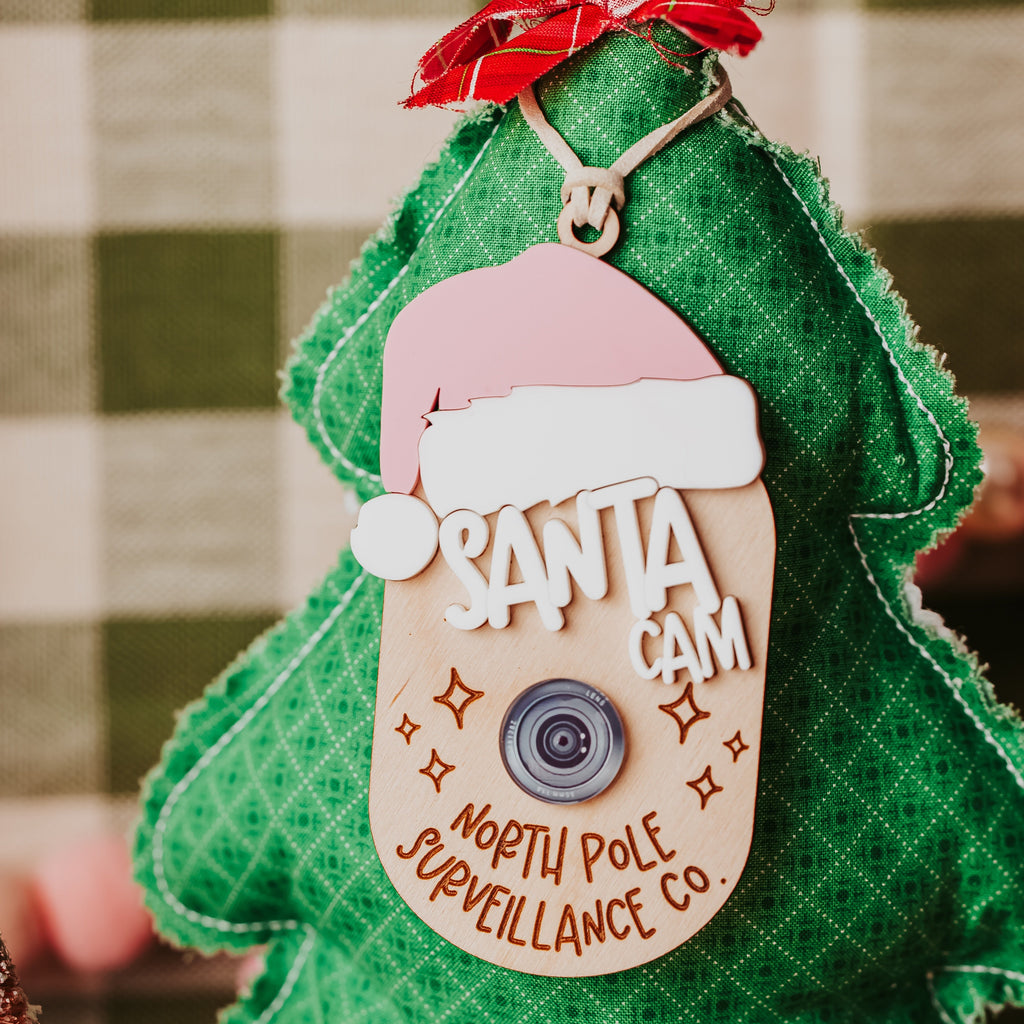 A green Christmas tree-shaped fabric decoration with a faux 'Sant Cam; ornamentreading 'SANTA CAM' and 'NORTH POLE SURVEILLANCE CO.', with a pink Santa hat.
