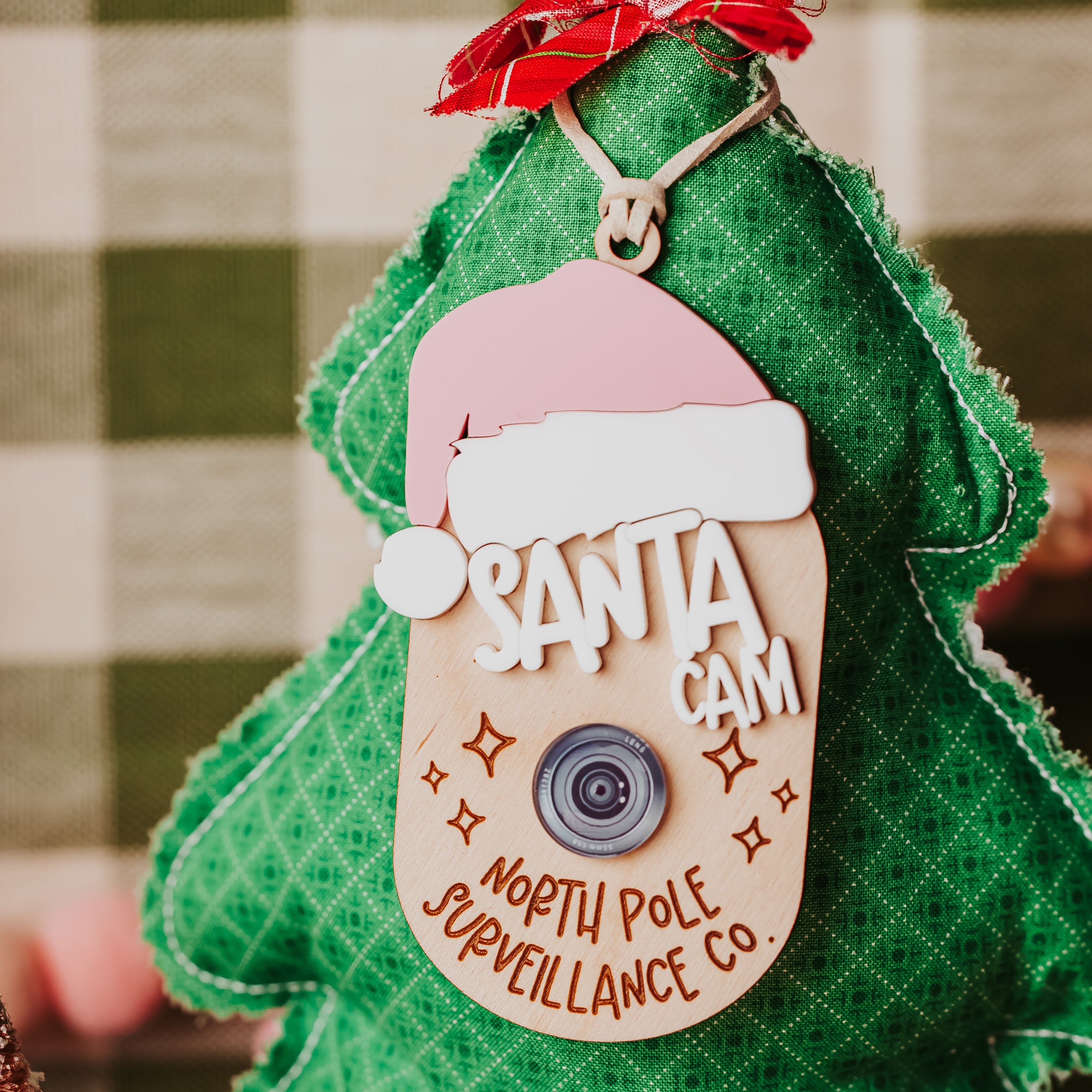 A green Christmas tree-shaped fabric decoration with a faux 'Sant Cam; ornamentreading 'SANTA CAM' and 'NORTH POLE SURVEILLANCE CO.', with a pink Santa hat.