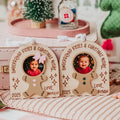 Two personalized gingerbread man photo ornaments displayed on a cozy holiday table setting with cookies, a pink suitcase, and Christmas decorations in the background.