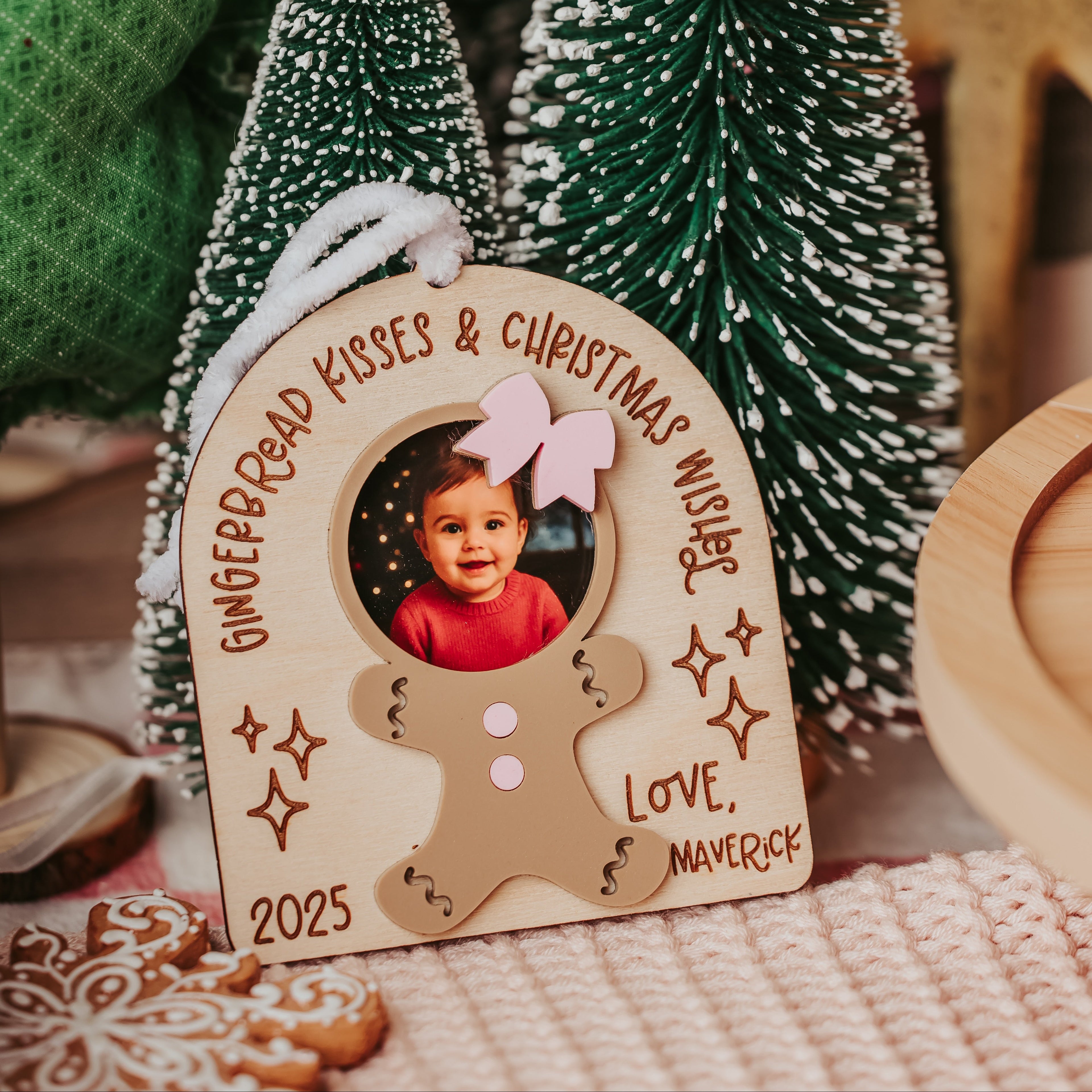 Personalized wooden Christmas ornament with a gingerbread man silhouette, custom photo frame, and engraved text reading 'Gingerbread Kisses & Christmas Muffins' and 'Love, Maverick 2025', styled with miniature Christmas trees and holiday decor.