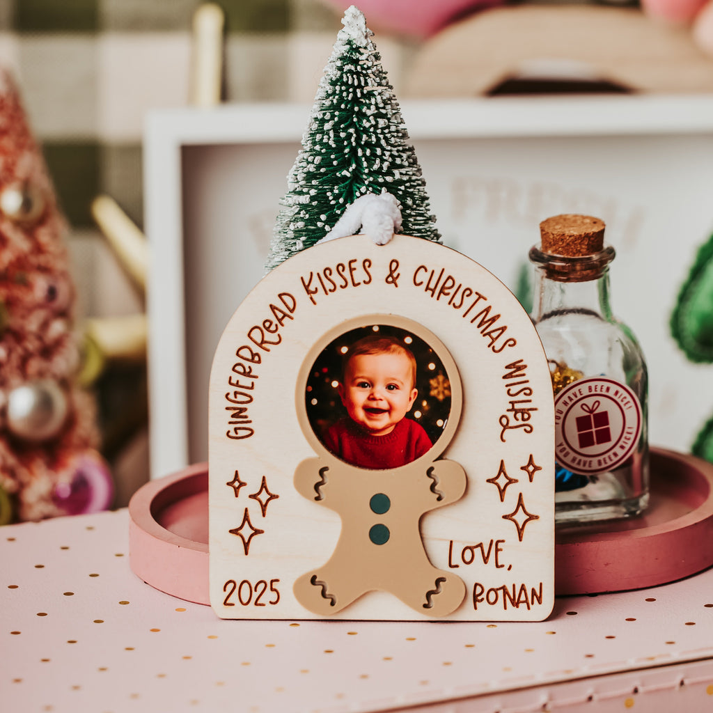 A personalized gingerbread cookie-shaped photo frame displayed on a pink polka-dot surface. The frame has a photo of a child in the center, with the text 'GINGERBREAD KISSES & CHRISTMAS WISHES' and 'LOVE, RONAN' around the top and bottom.