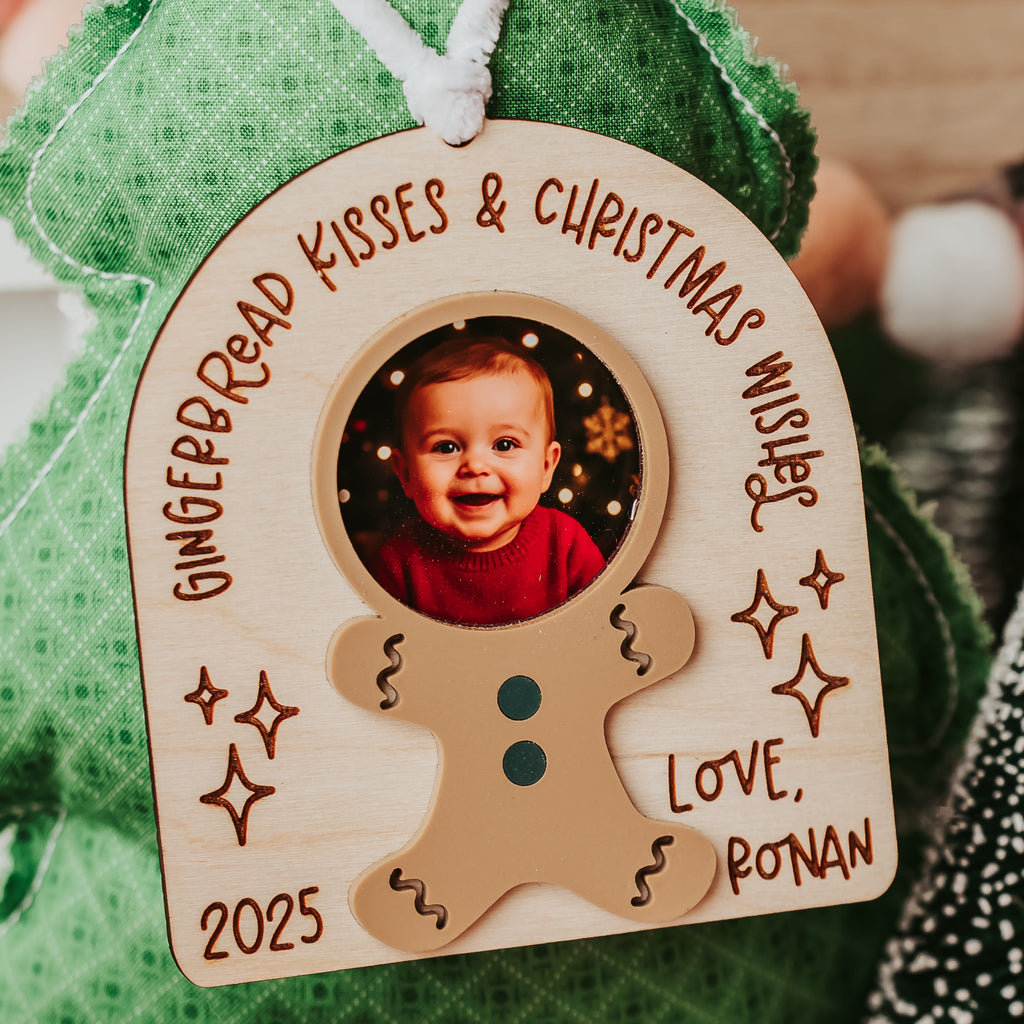 A personalized wooden Christmas ornament shaped like a gingerbread man, featuring a photo of a smiling child, the text 'GINGERBREAD KISSES & CHRISTMAS WISHTS', 'LOVE, RONAN', and '2025'. The ornament is hanging from a green Christmas tree decoration with a red bow.
