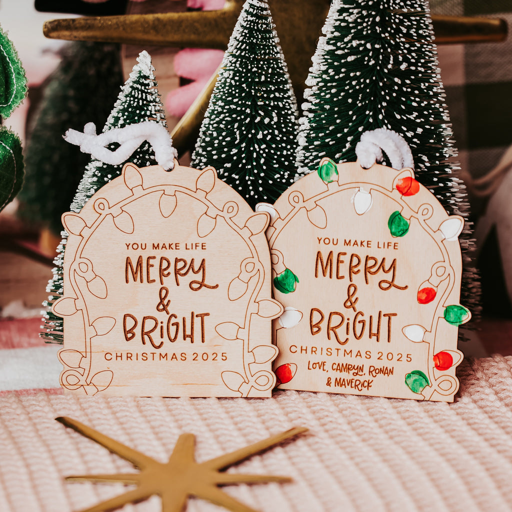Two decorative arch-shaped wooden Christmas ornaments hang on a table, each printed with the phrase 'YOU MAKE LIFE MERRY & BRIGHT CHRISTMAS 2025' in brown lettering. The ornaments are styled with small green and white miniature Christmas trees, a gold star, and a pink knit blanket in the background.